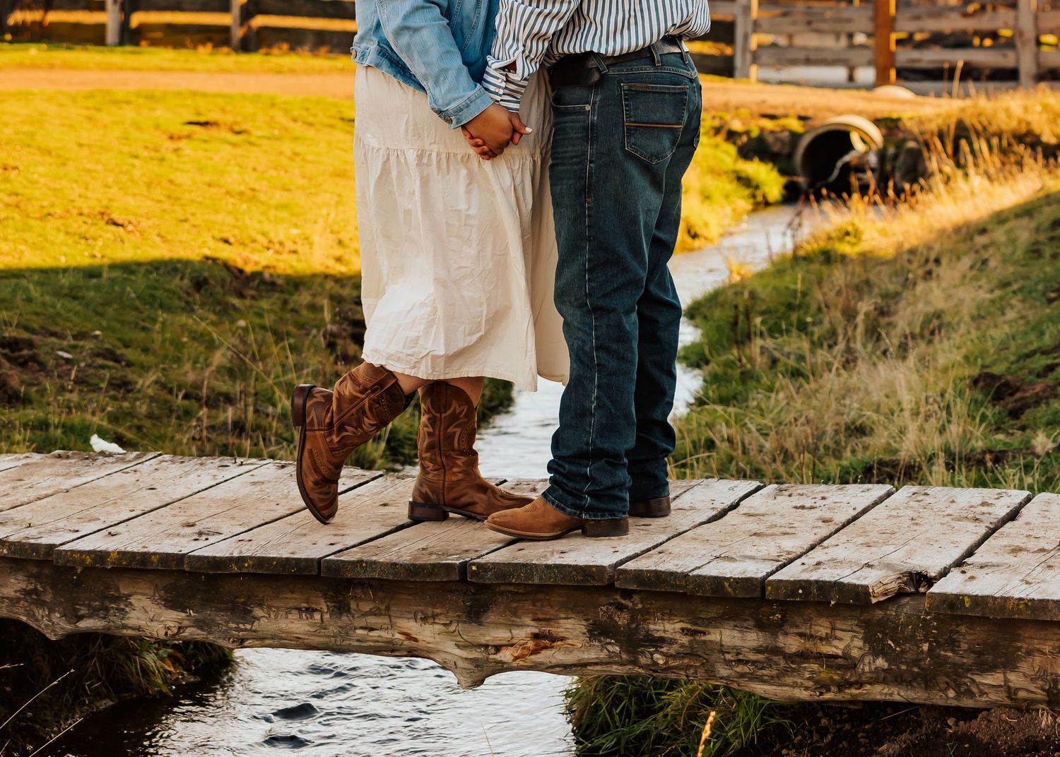 A couple stands on wooden planks near a stream during a countryside photoshoot.