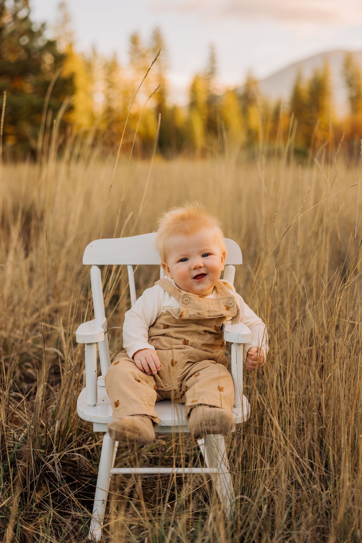 Smiling baby in beige outfit sits in white chair among tall grass at sunset.