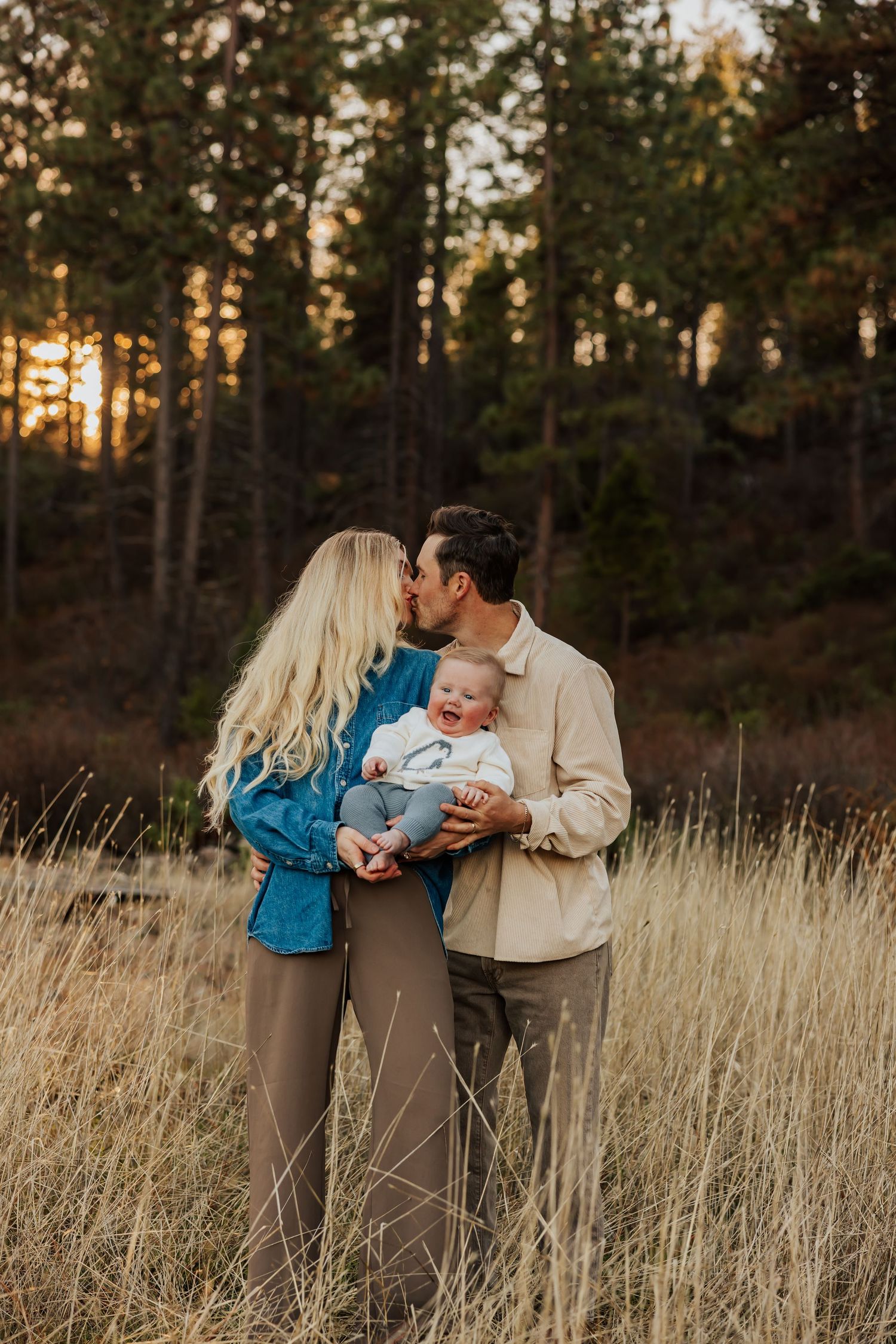 Couple in casual attire shares tender moment with baby in field during golden hour.