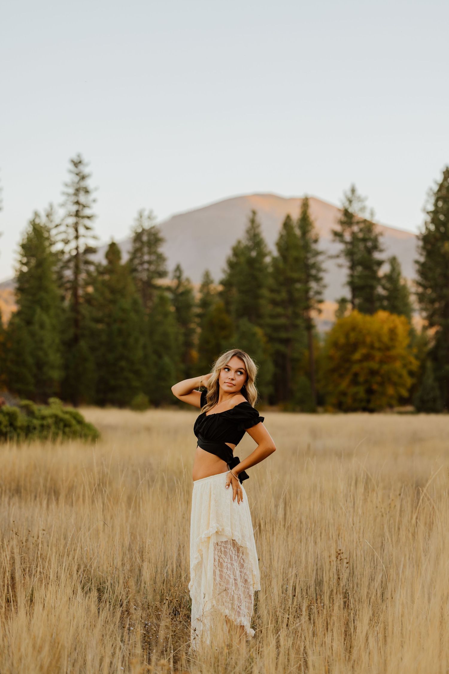 Outdoor portrait session in a golden wheat field with mountains and evergreen trees in the background.