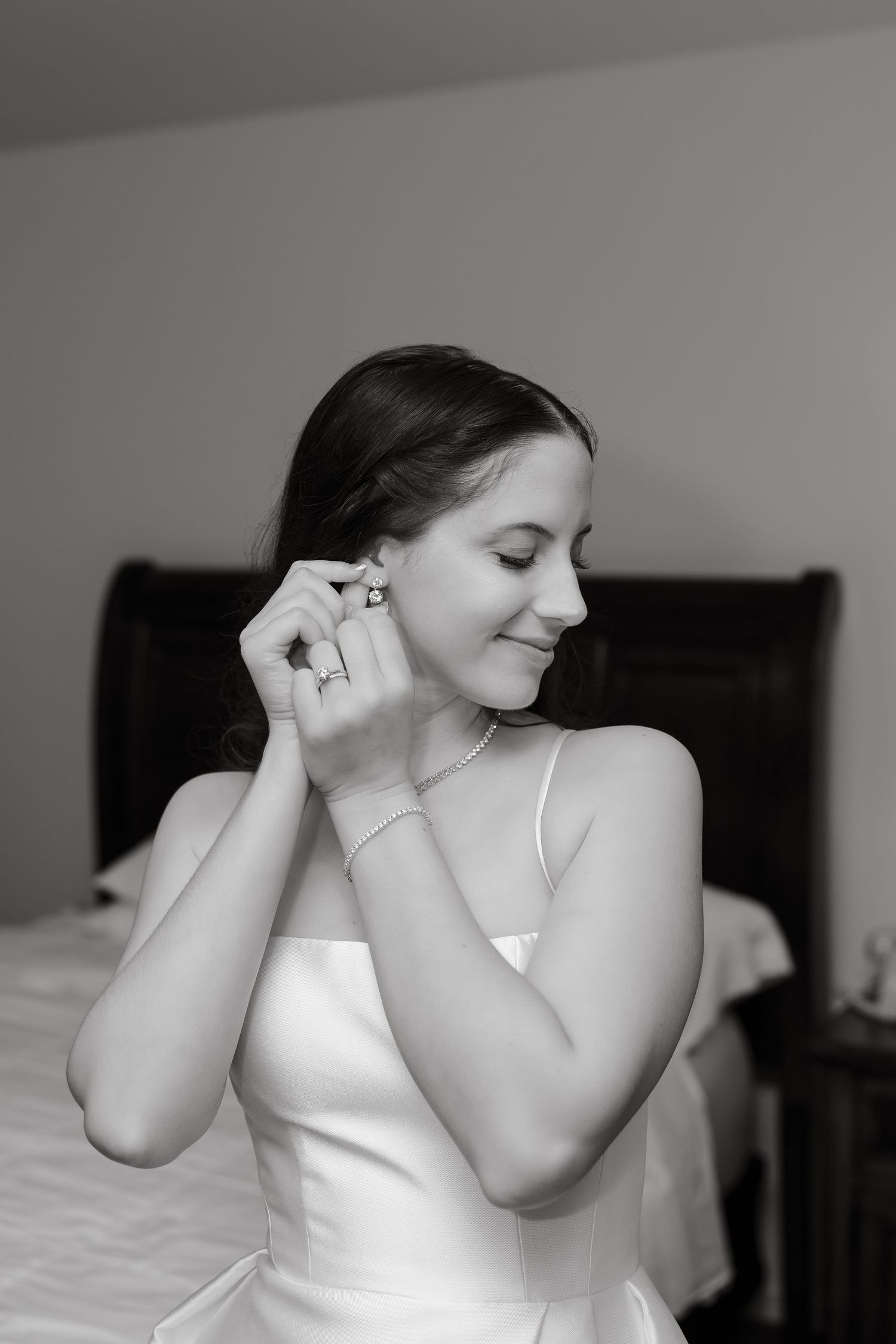 Bride in white dress putting on earrings while smiling, black and white wedding preparation photo.