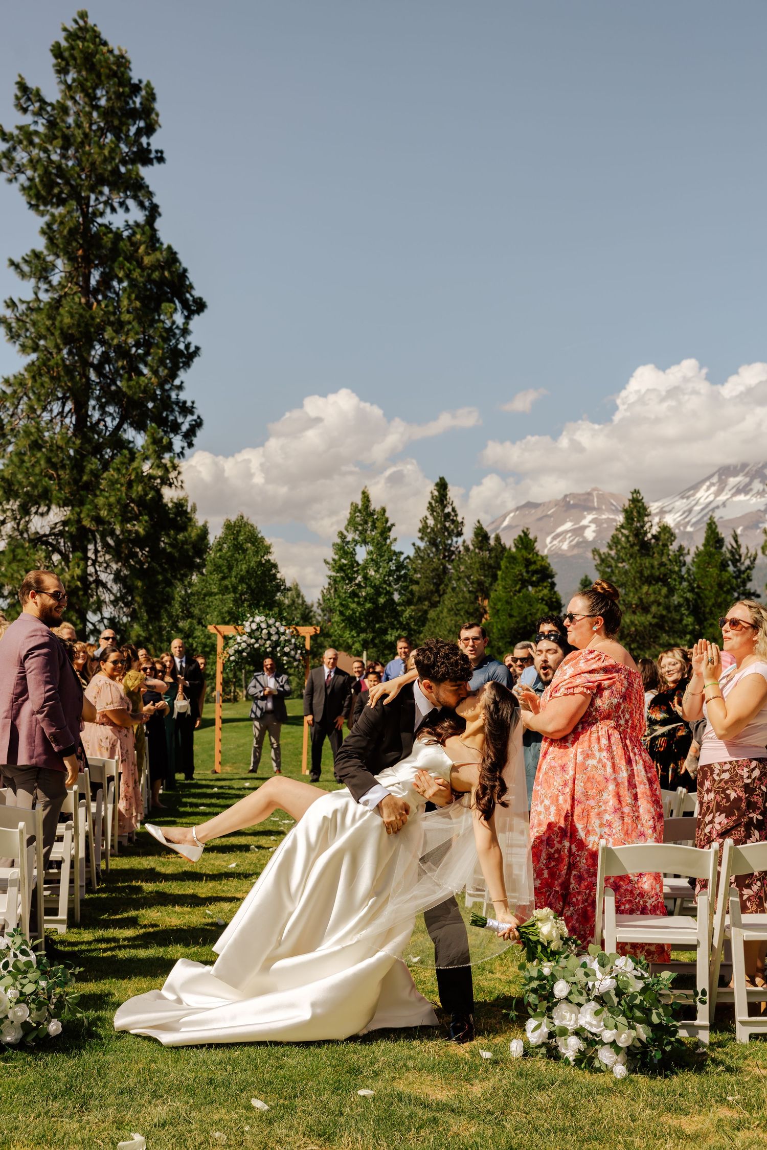 Groom dips bride in romantic kiss along outdoor wedding aisle with snowy mountain views.