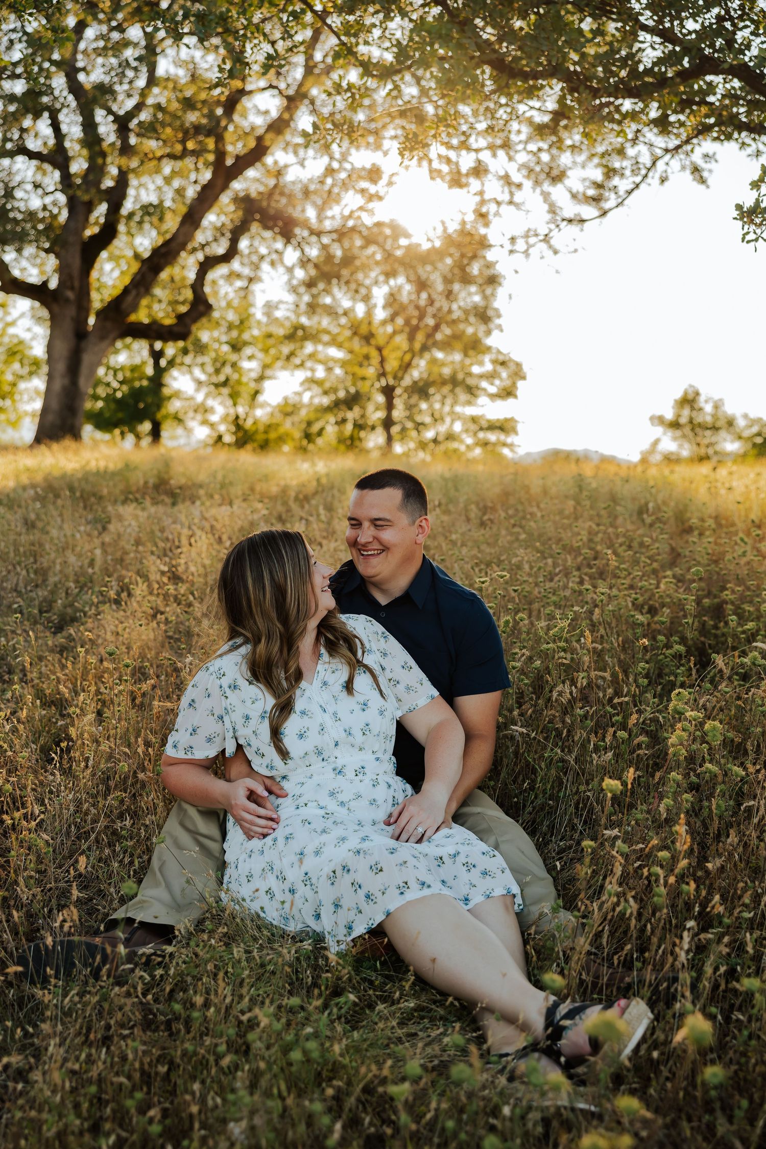 Couple sitting in golden field at sunset, woman in floral dress and man in navy polo sharing a romantic moment under oak trees.