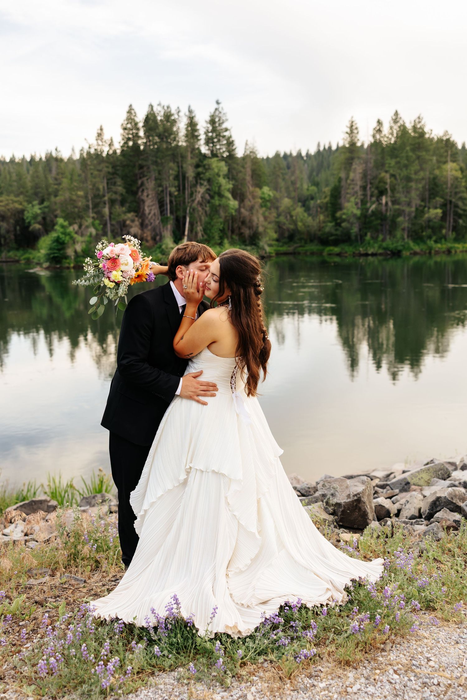 Bride and groom share a romantic kiss by a serene lake surrounded by evergreen trees, bride holding a colorful bouquet.