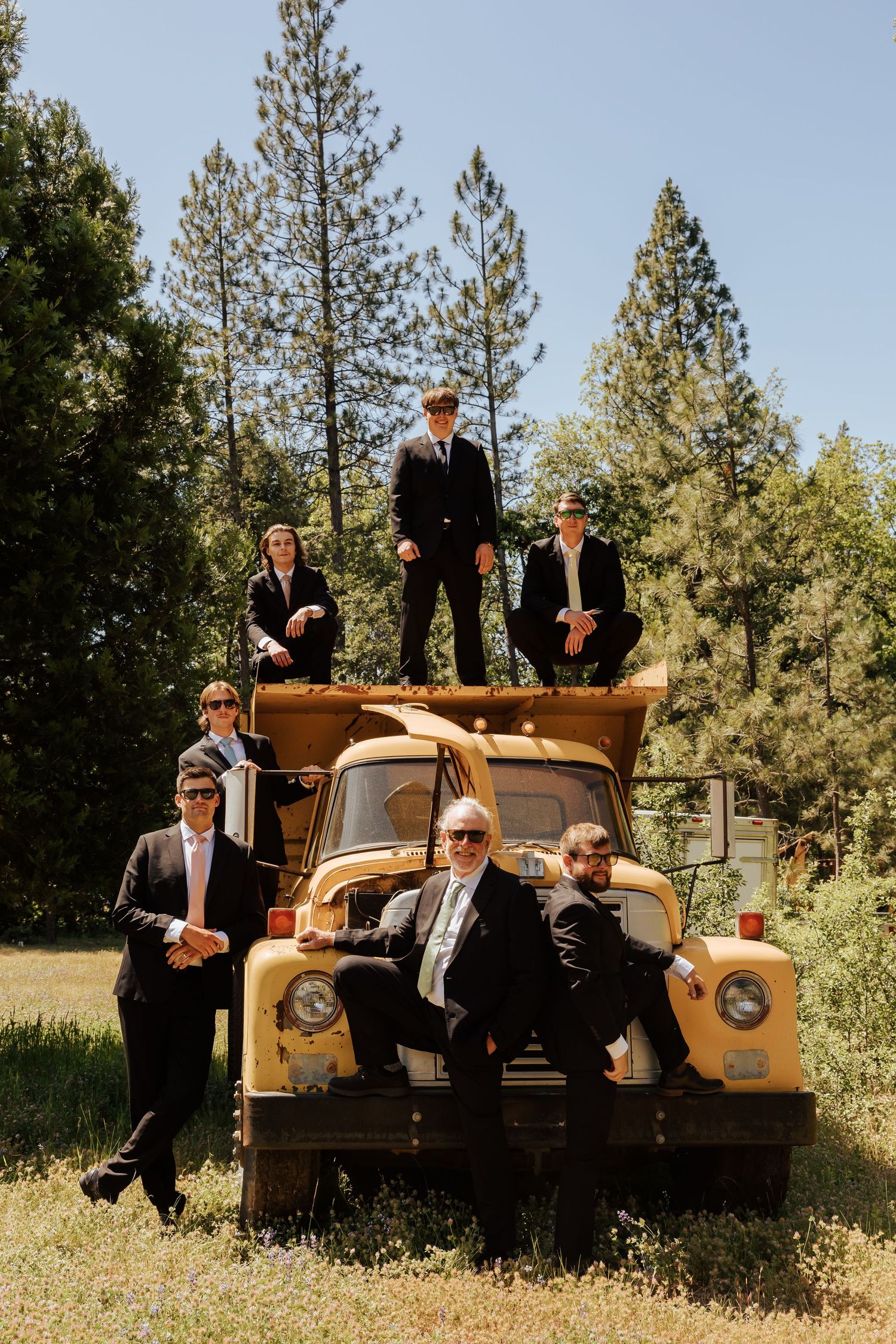 Groomsmen in black suits pose on a vintage yellow truck surrounded by tall pine trees on a sunny wedding day.