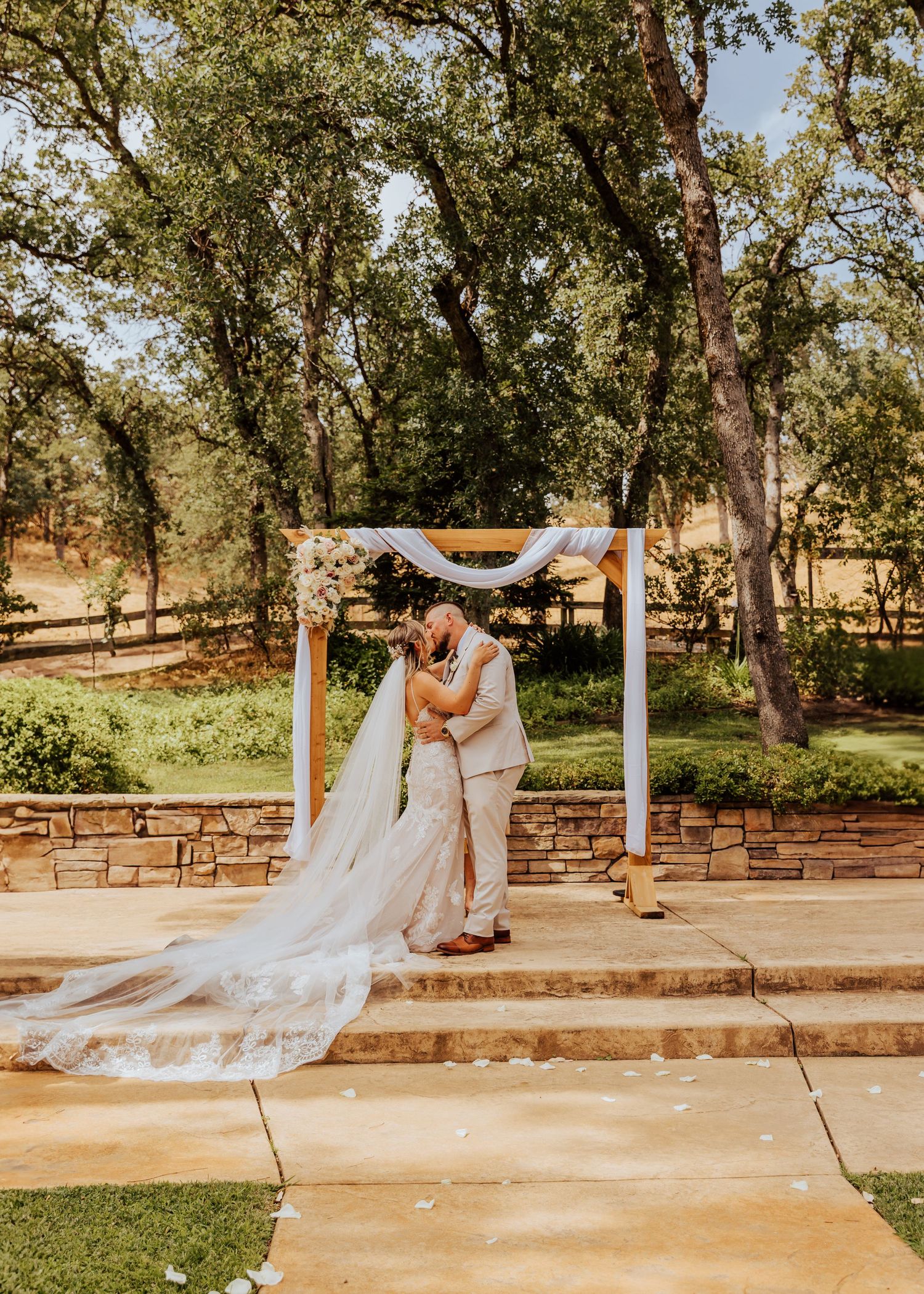 Romantic outdoor wedding ceremony under trees with white drapery arch and stone steps.