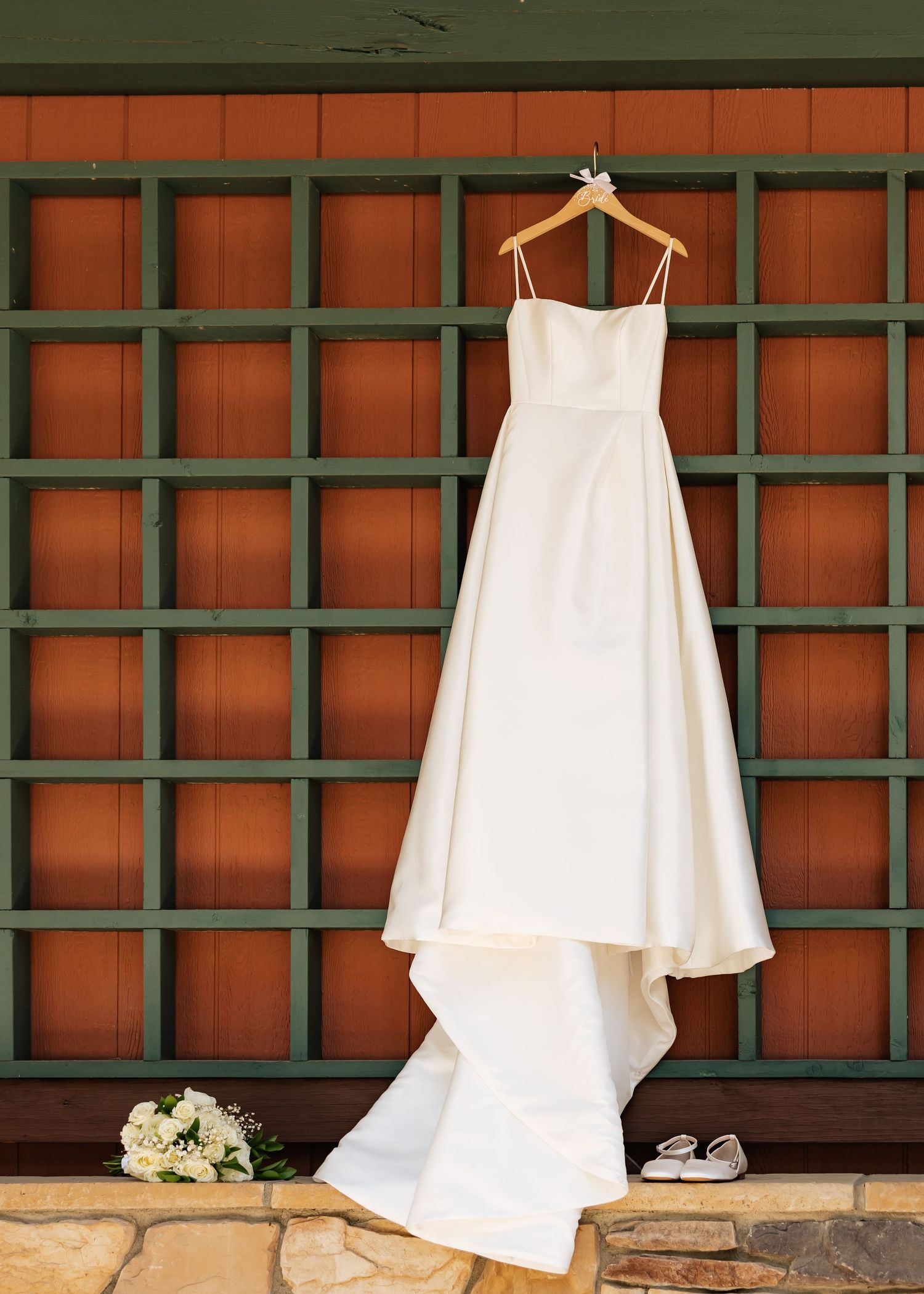 White satin wedding gown with spaghetti straps hanging on wooden hanger against rustic red and green grid wall.