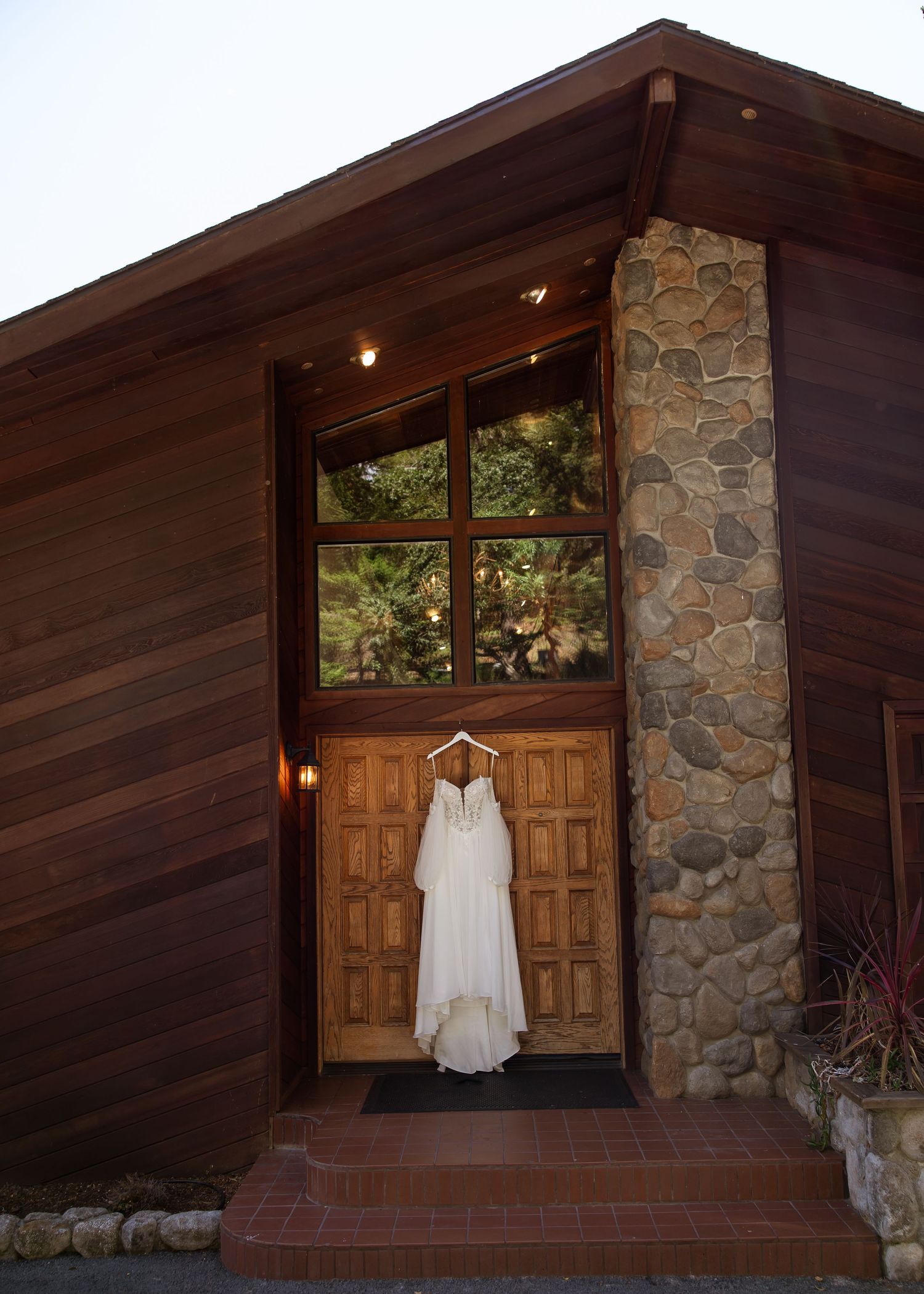 A white wedding dress hanging on wooden double doors of a rustic stone and wood chapel venue.