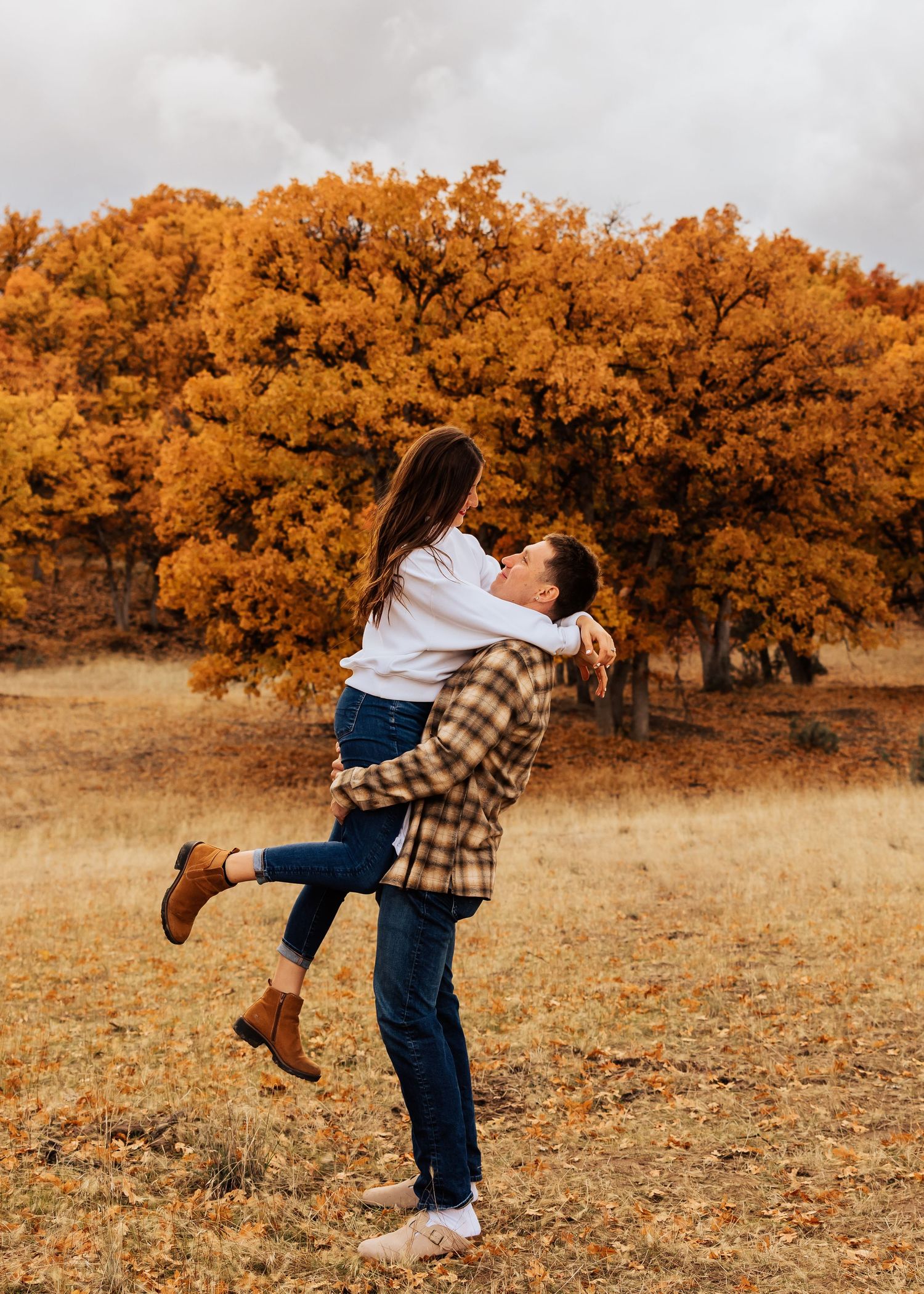 Playful couple spinning and embracing in a field with orange autumn trees in the background.