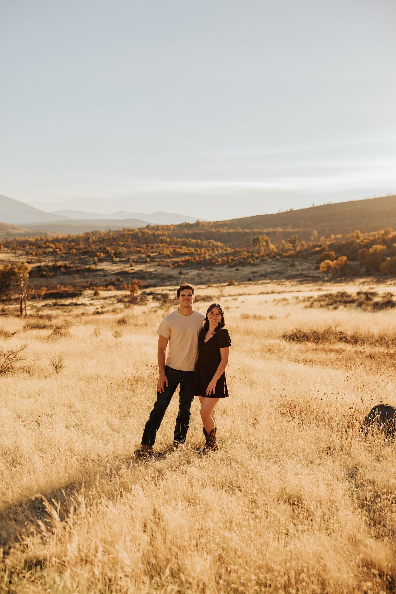 Couple sharing romantic moments in a golden sunset field with mountains in background.