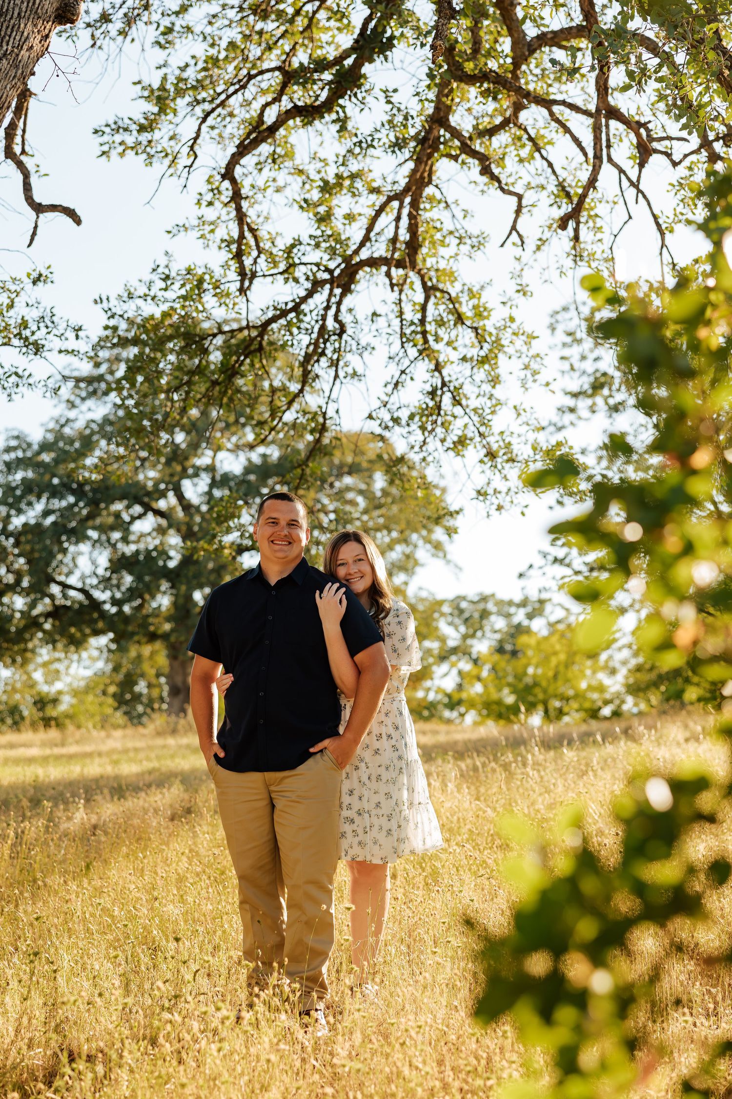 Smiling couple standing in sunlit meadow under large oak tree branches.