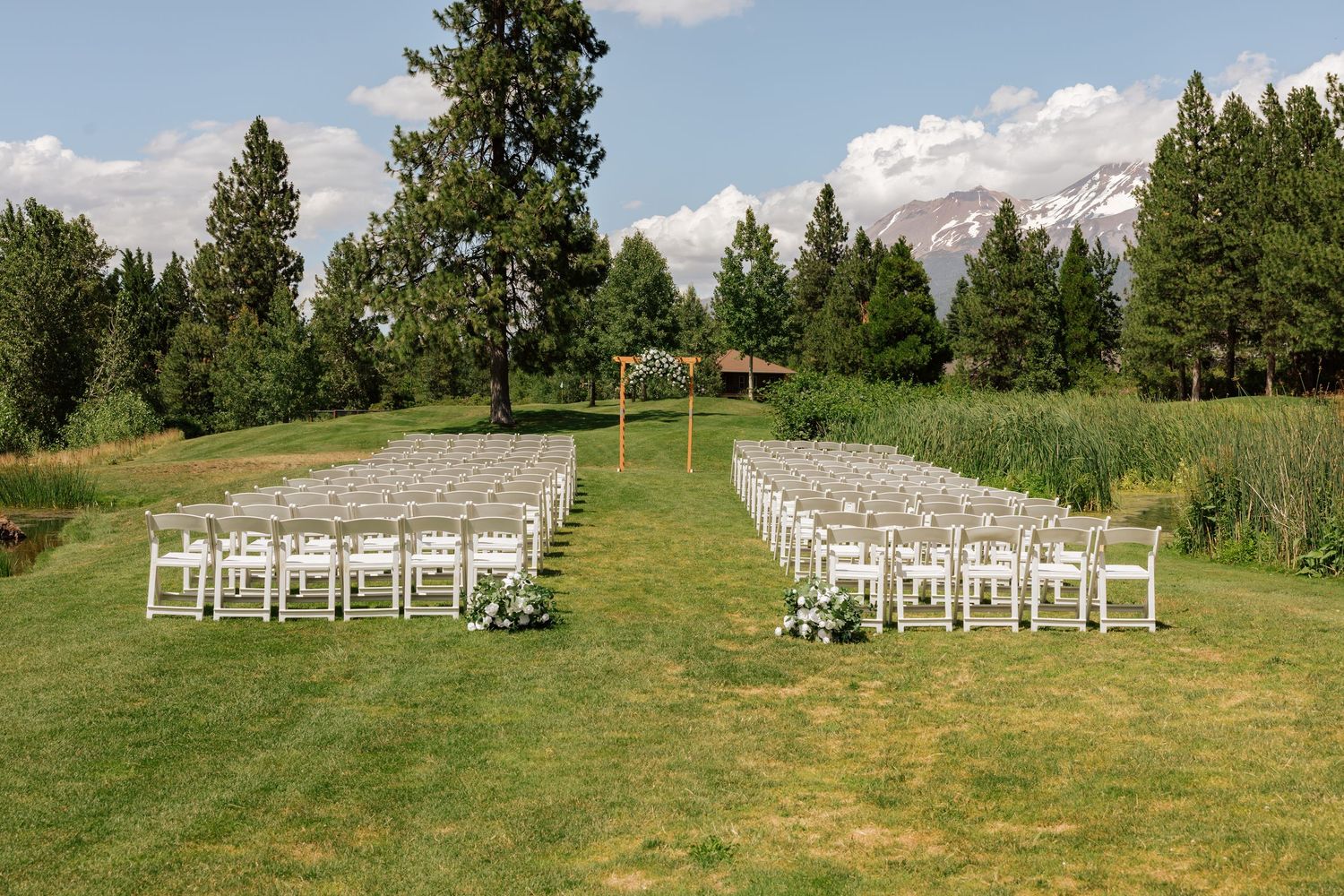 Outdoor wedding ceremony setup with white chairs on green lawn, wooden arch, tall pines, and snow-capped mountains in background.