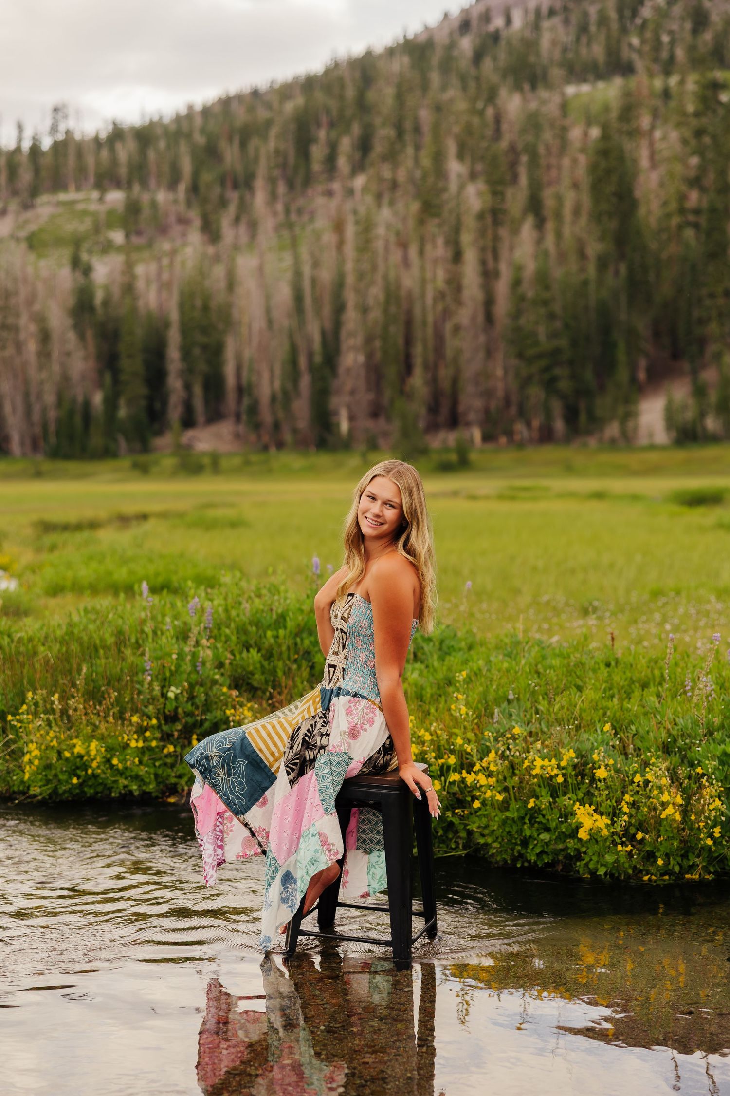 Young blonde woman in colorful patchwork dress sitting on stool in mountain stream with green meadow backdrop.