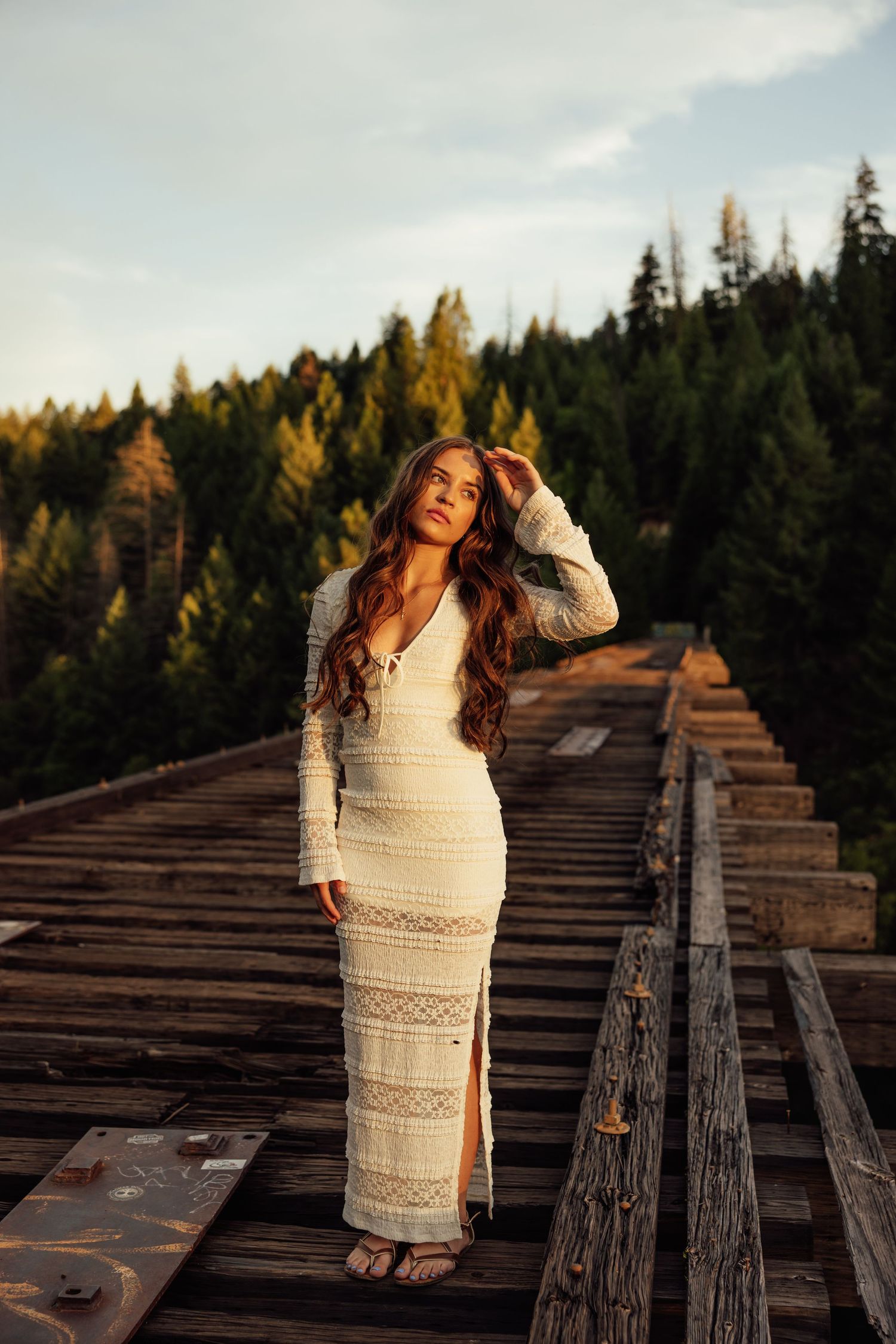 A person in a white crochet dress poses on wooden railroad tracks with forest scenery.