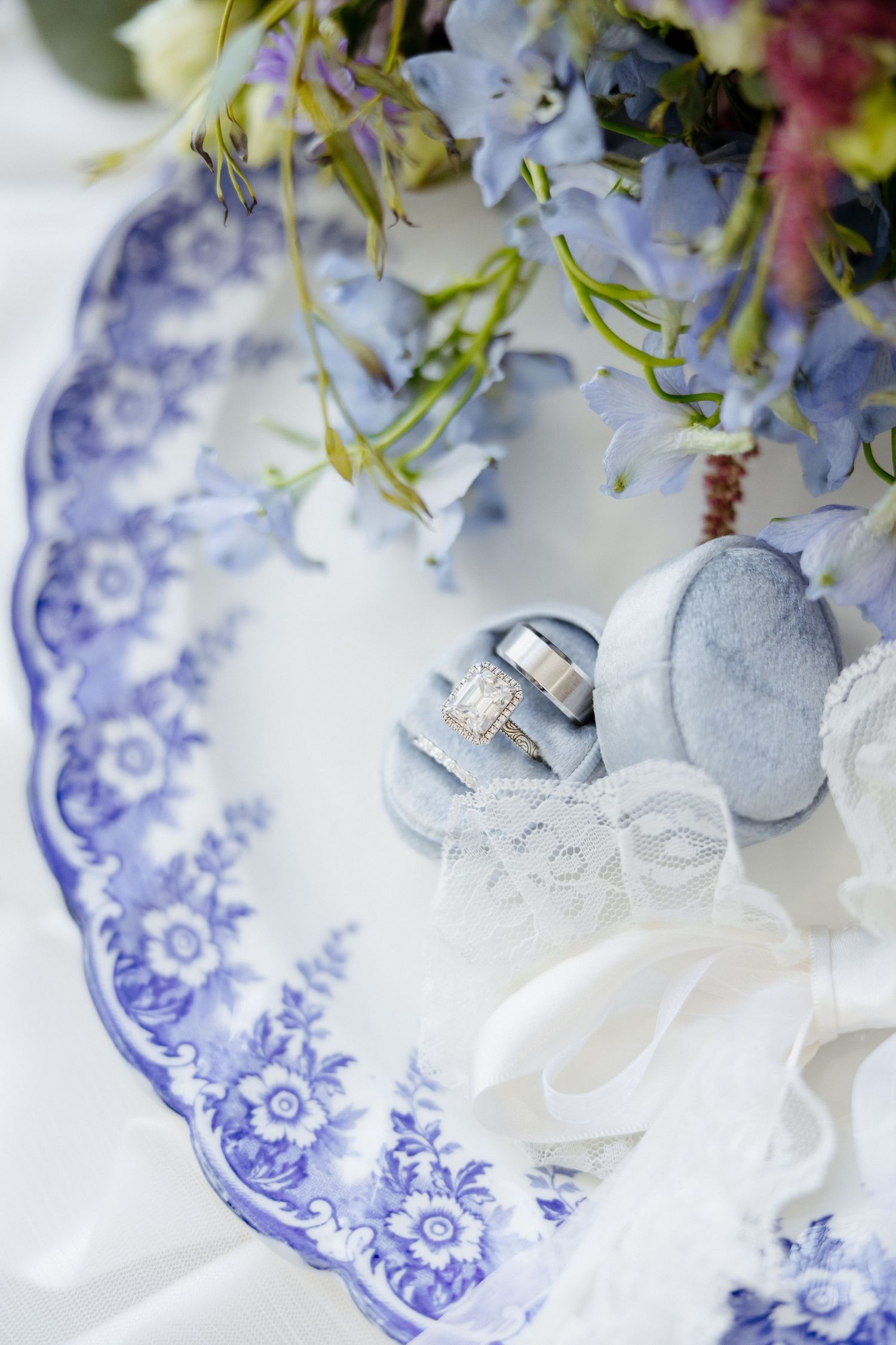 Blue and white floral plate with wedding rings in velvet box, lace ribbon, and blue flowers bouquet.