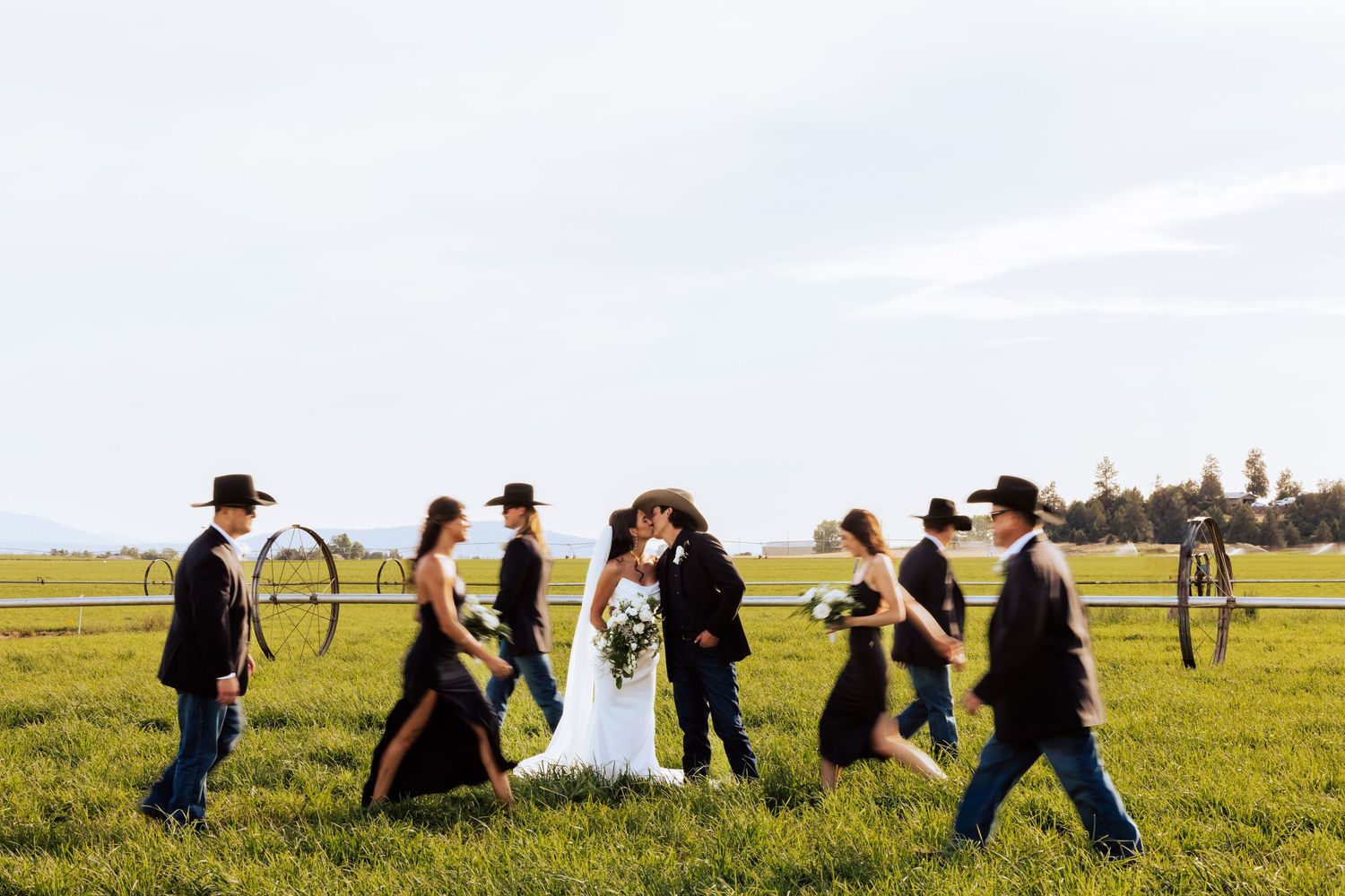 Wedding party walks through green field, bride and groom kiss, groomsmen in cowboy hats carry lasso hoops.