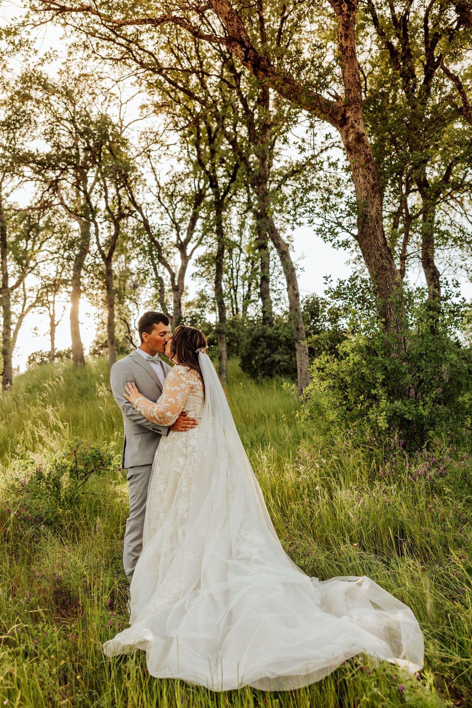 Bride and groom share a kiss in sunlit forest meadow, wedding dress train flowing through tall green grass.