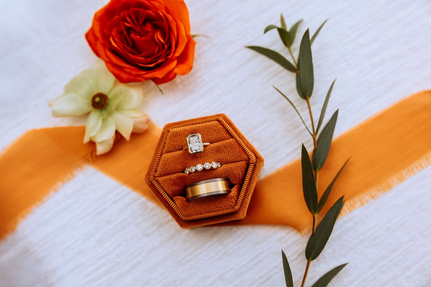 Wedding rings displayed with red roses and white flowers on an orange fabric background in a romantic flat lay arrangement.