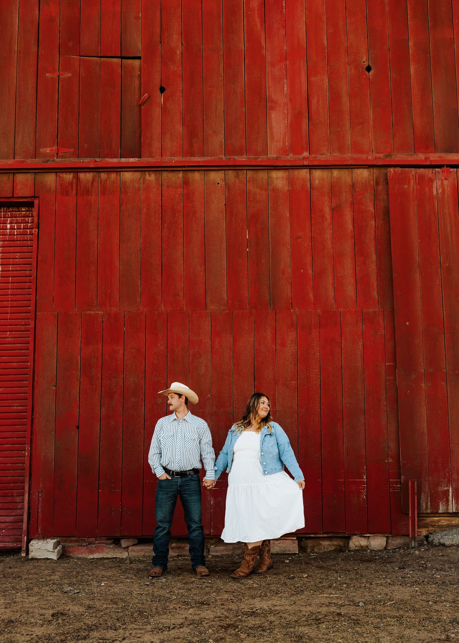 Couple in western wear holding hands against bright red barn wall.