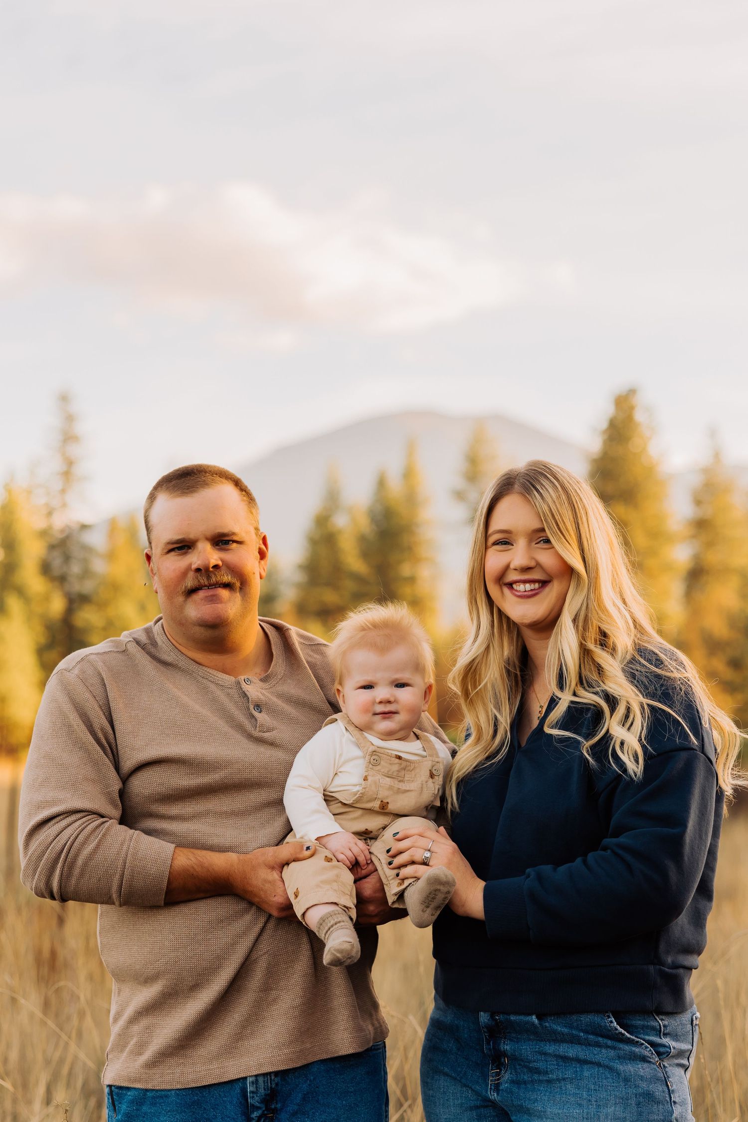Family portrait in a golden field with mountains and evergreen trees in background.