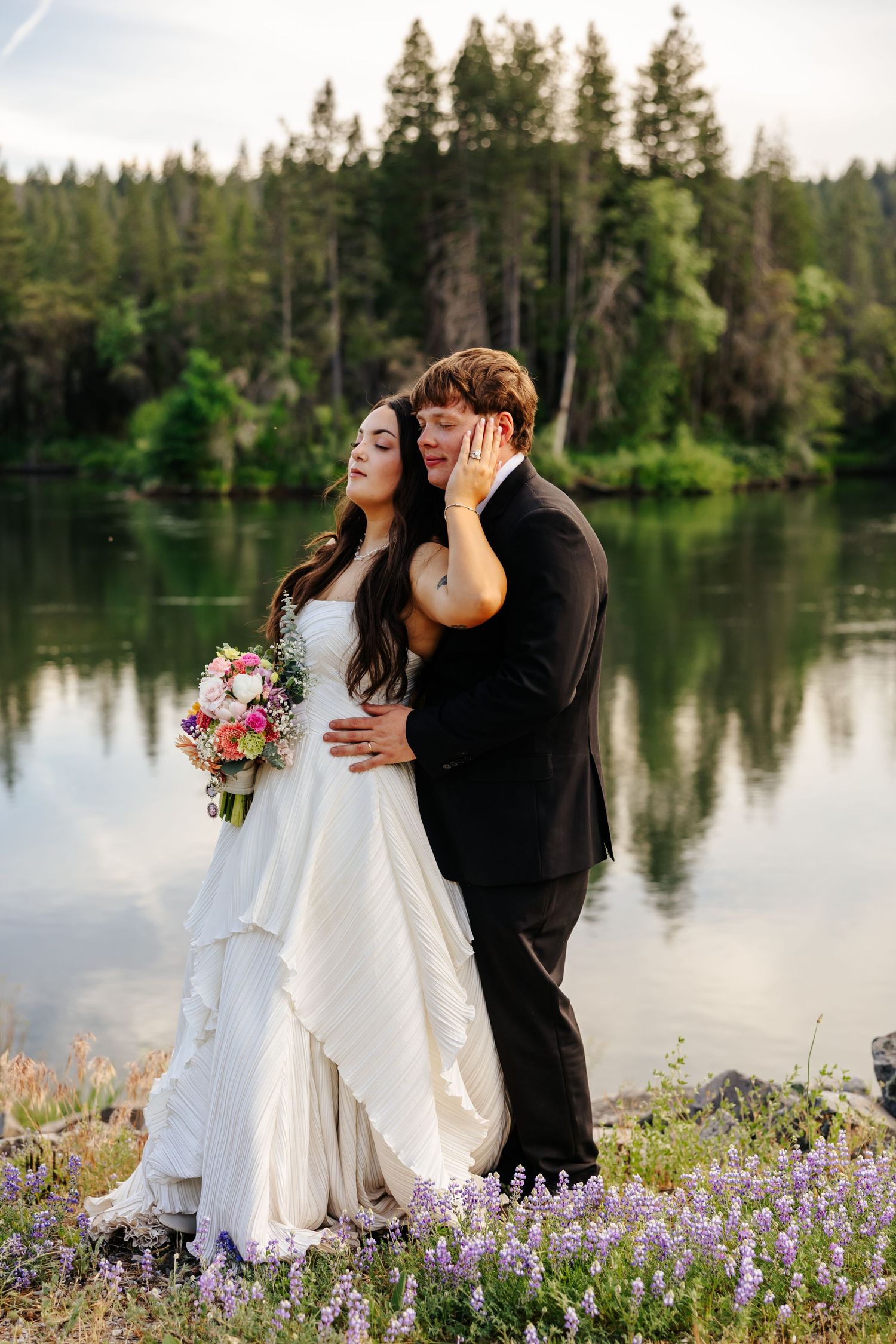 Bride and groom pose romantically by a reflective lake with purple wildflowers and pine forest backdrop.