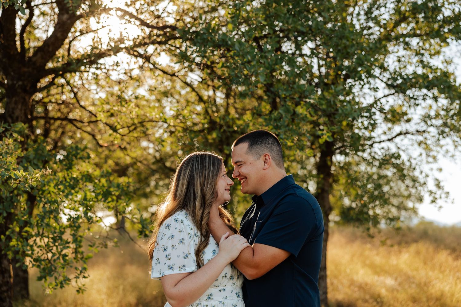 A couple shares a romantic moment outdoors, smiling at each other under golden sunlit trees in a field.
