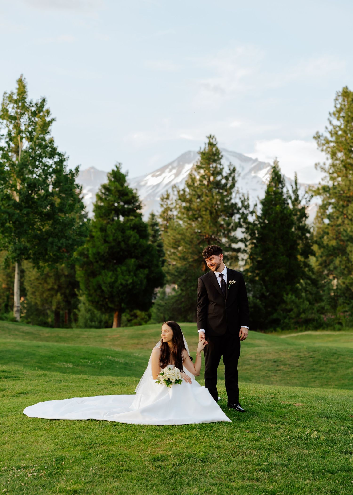 Bride in white gown sits gracefully on green lawn as groom stands beside mountain scenery.