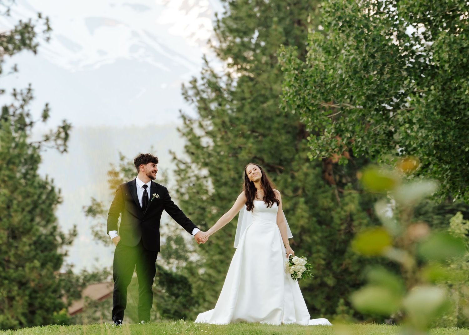 Bride and groom hold hands in a lush green garden with tall trees and mountain views on their wedding day.
