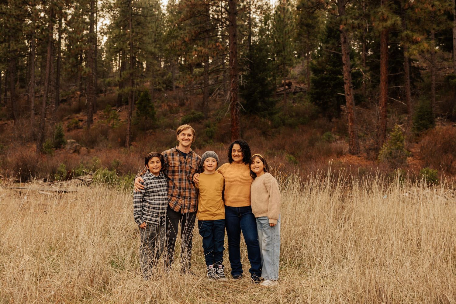 Family of four poses together in tall grass with forest backdrop during autumn.