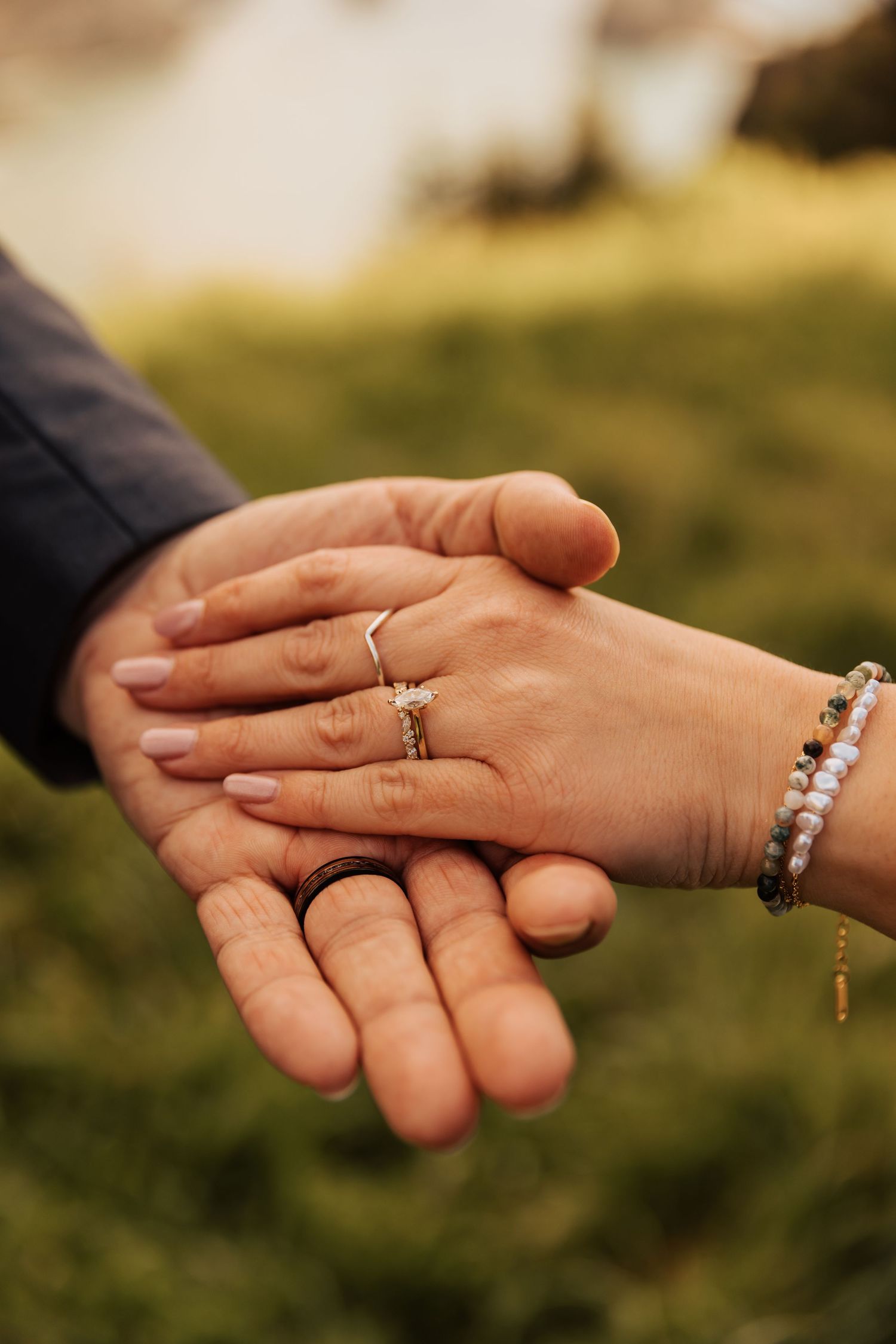 Close-up view of hands holding each other with engagement rings against a blurred outdoor background.