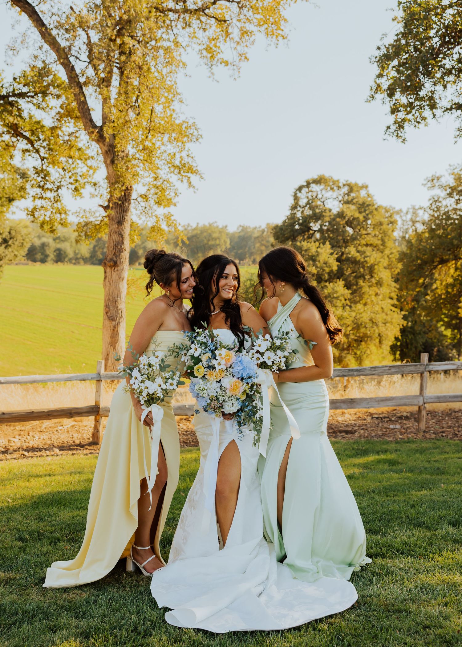 Bridesmaids in light blue dresses gather on a grassy hillside with bouquets.