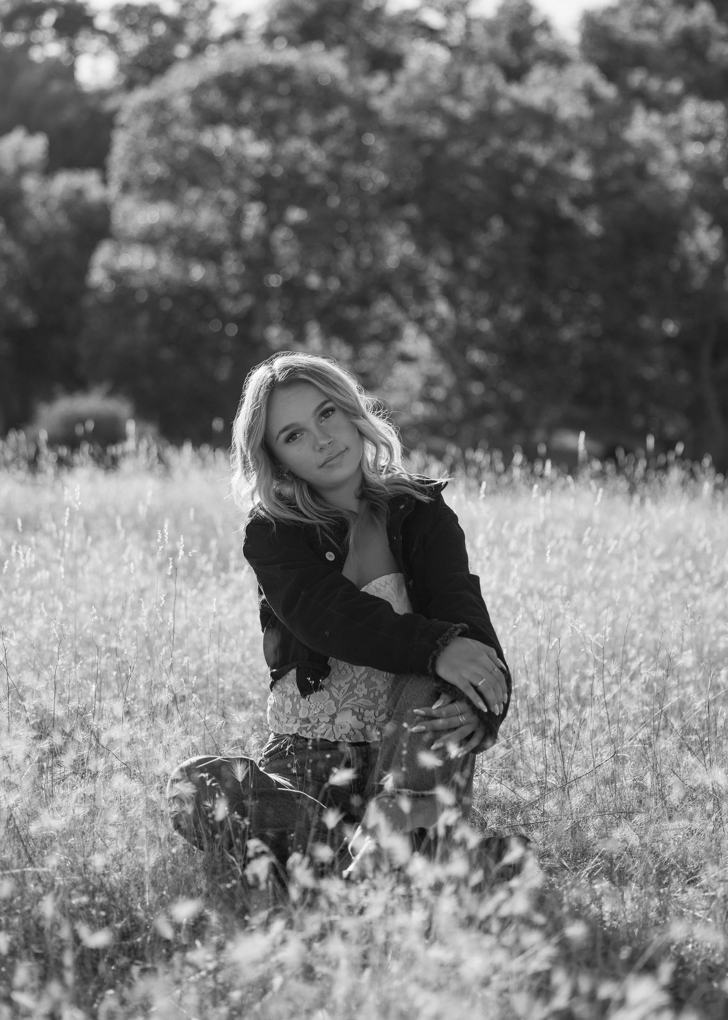 Black and white portrait of someone sitting in a wildflower meadow wearing a dark jacket and floral dress.