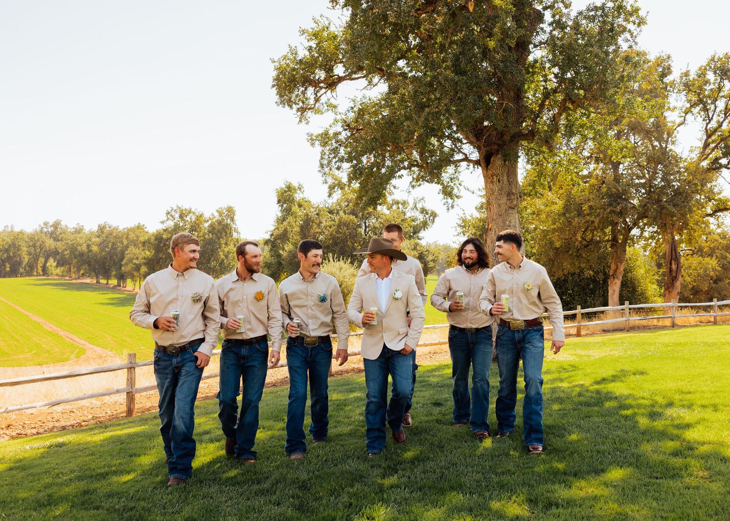 Wedding party in matching beige jackets and jeans stands in line on grassy field at sunset.