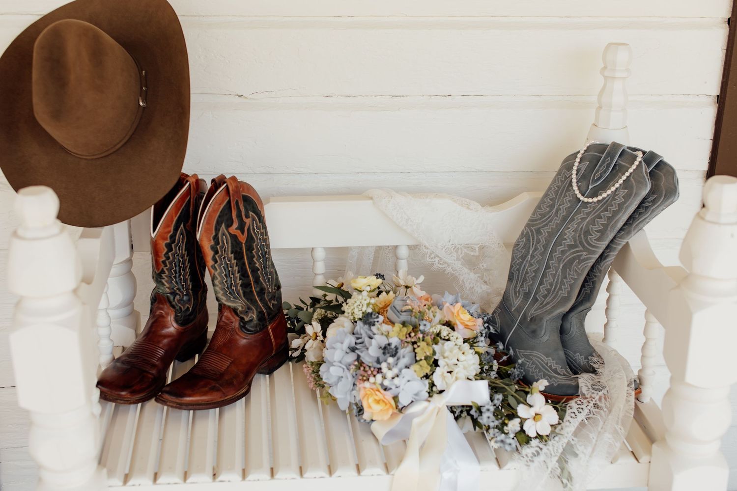 Wedding accessories including cowboy boots, a brown hat, and delicate floral bouquets arranged on a white bench.