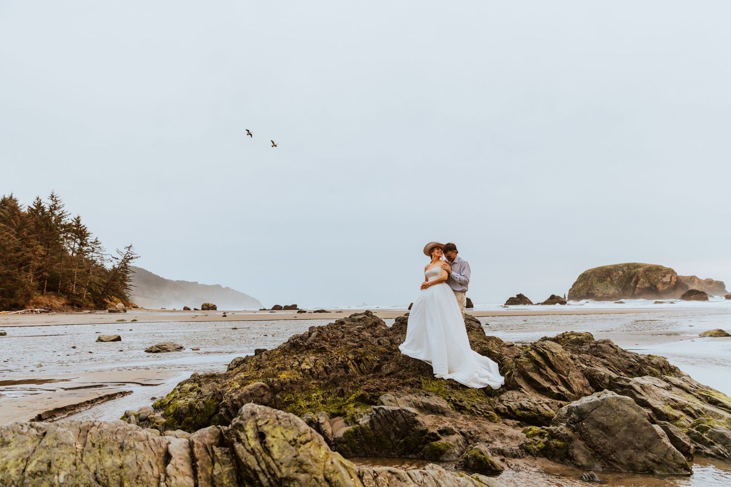 Wedding couple poses on dramatic rocky coastline with crashing waves in background.