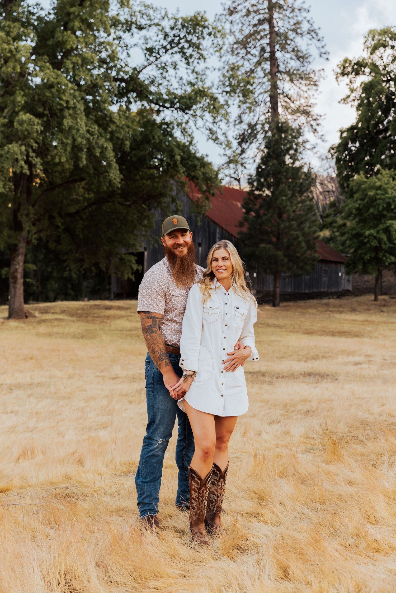 A couple stands together in a golden wheat field during sunset wearing western-inspired outfits.