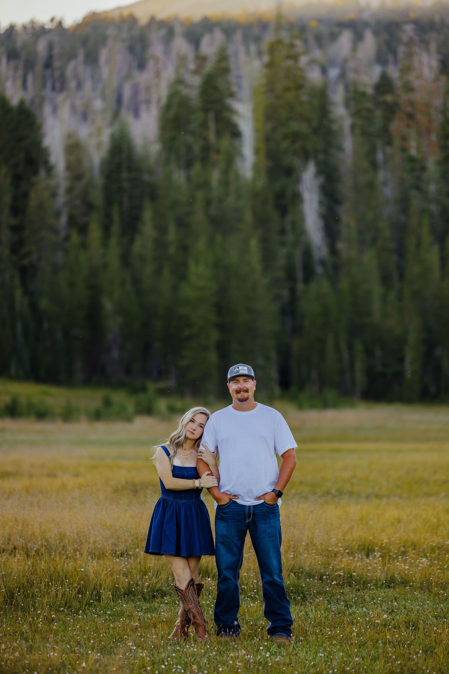 Couple posing in a meadow with tall pine trees and rocky mountains in the background during golden hour.