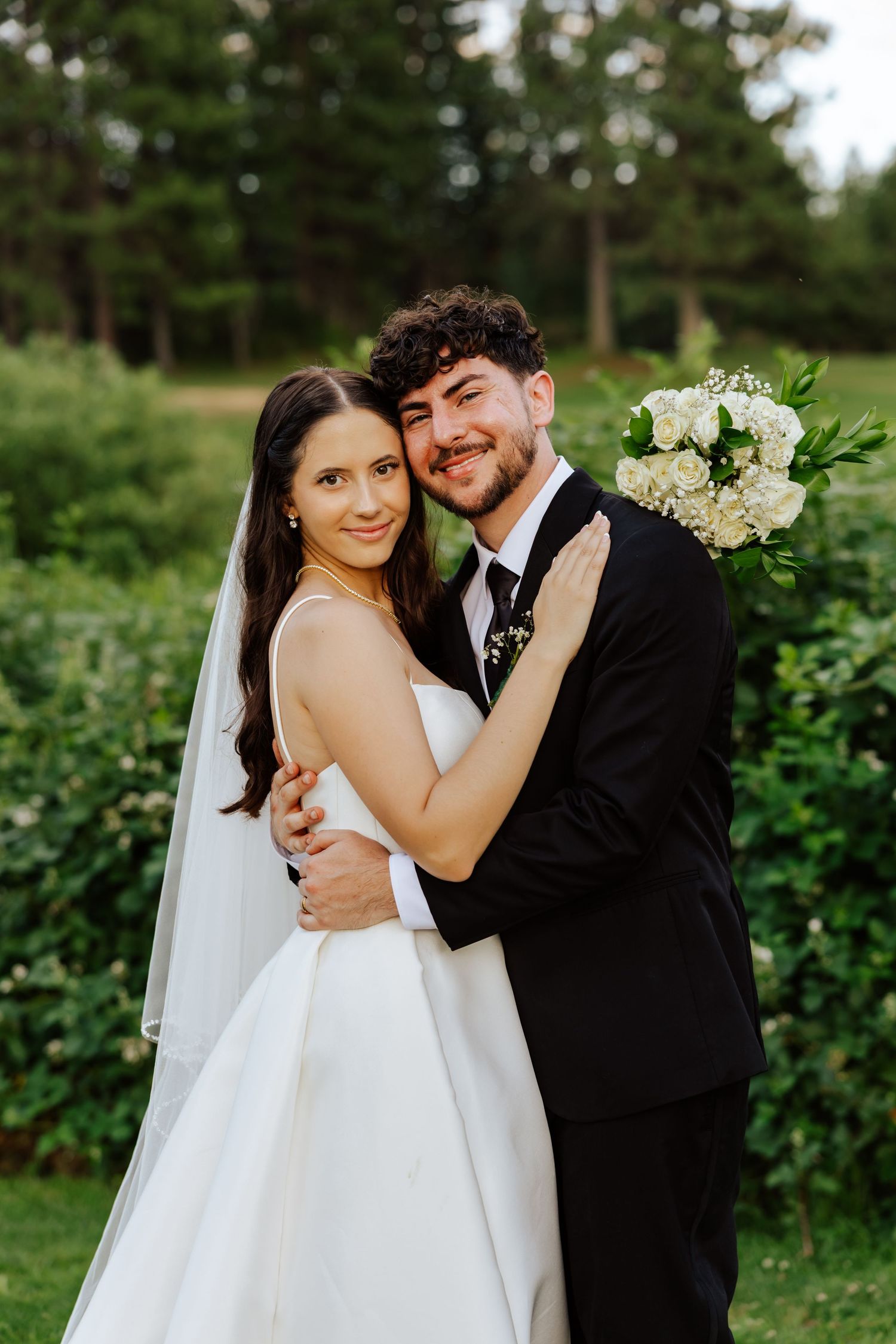 A bride in a white gown and veil embraces a groom in a black suit holding white flowers outdoors.