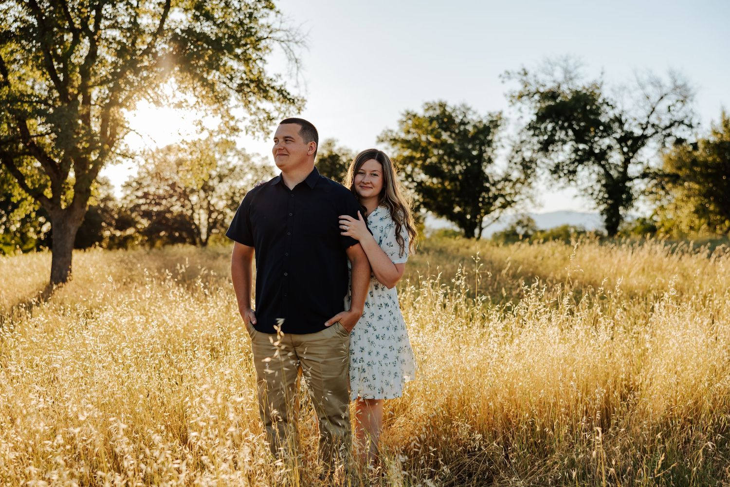 Couple standing together in a golden meadow at sunset, woman in floral dress embracing man in black shirt.