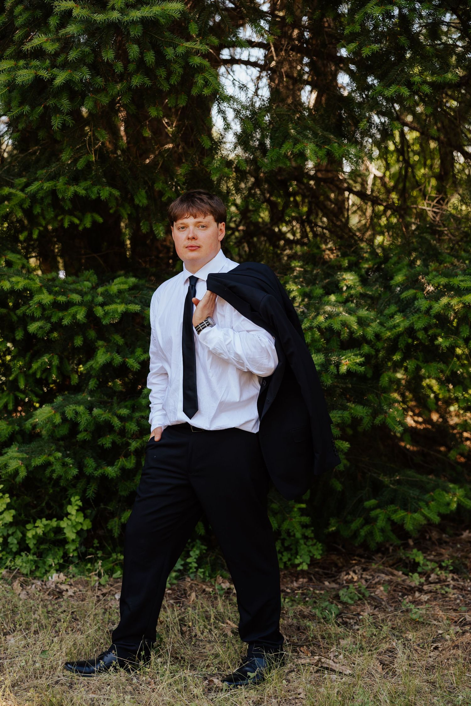Groom in white shirt and black tie carries suit jacket over shoulder while posing among green pine trees outdoors.