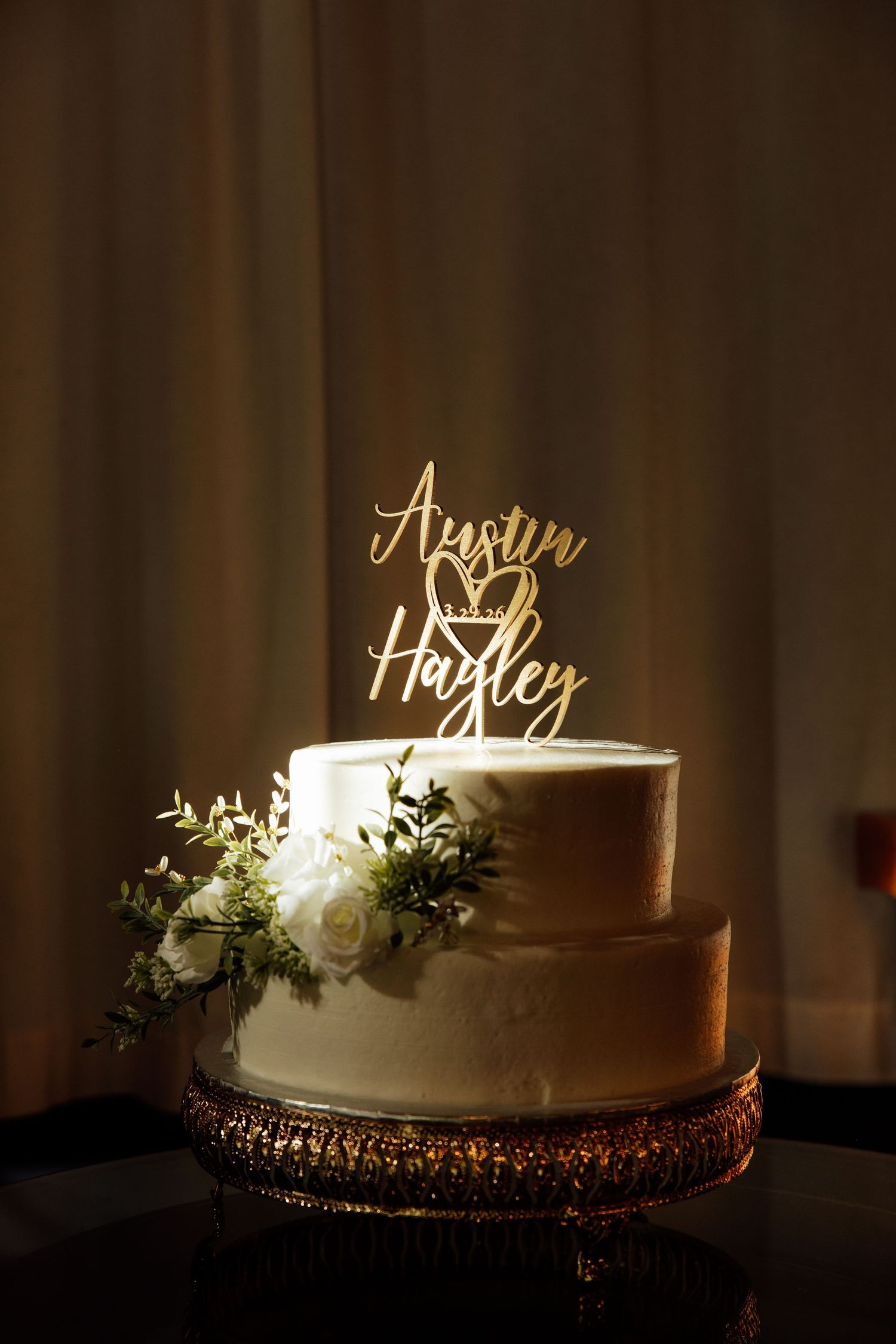 Elegant two-tier white wedding cake with gold topper reading Austin and Hayley, adorned with white roses and greenery.