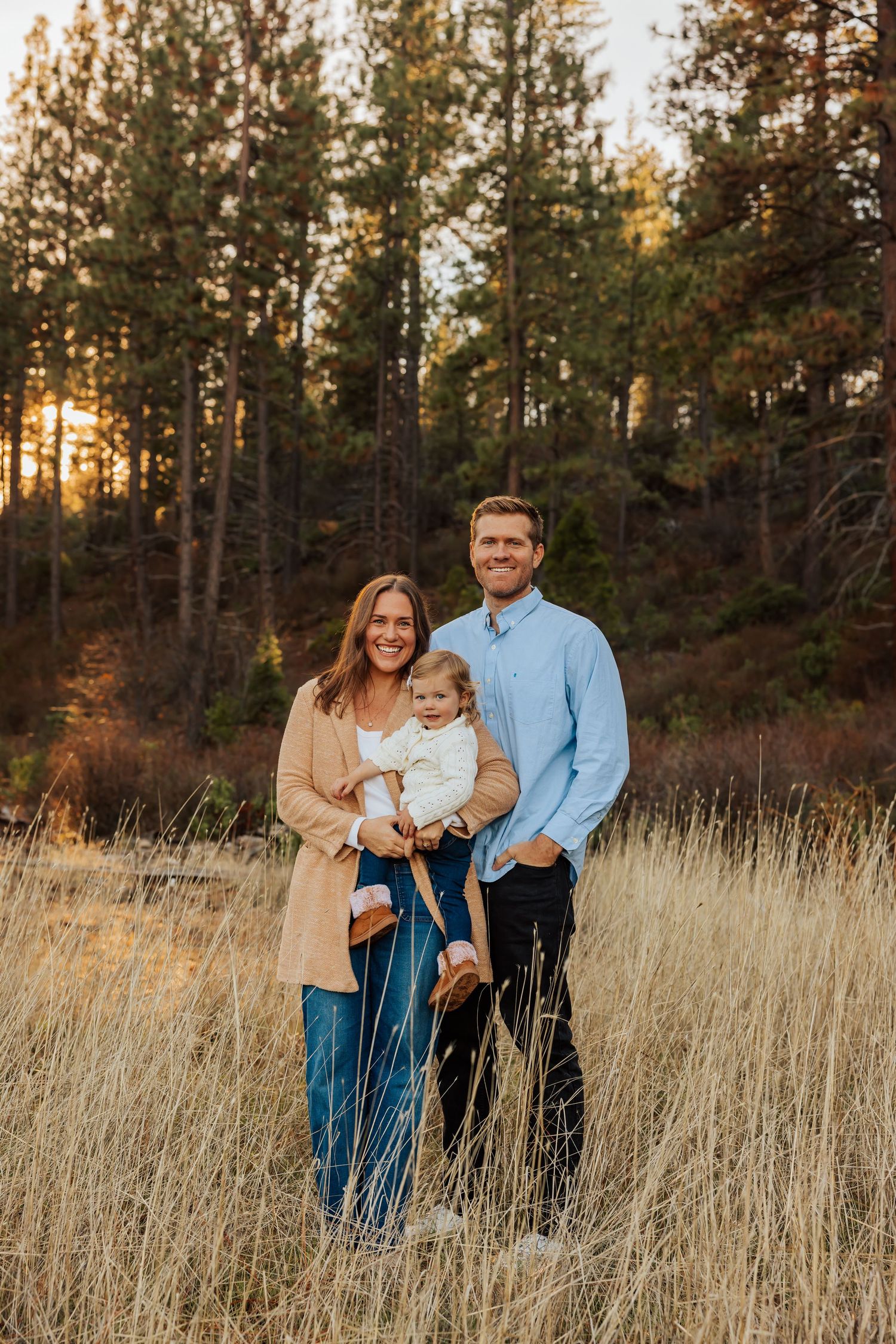 Small family poses together in casual attire against tall pine trees at sunset.