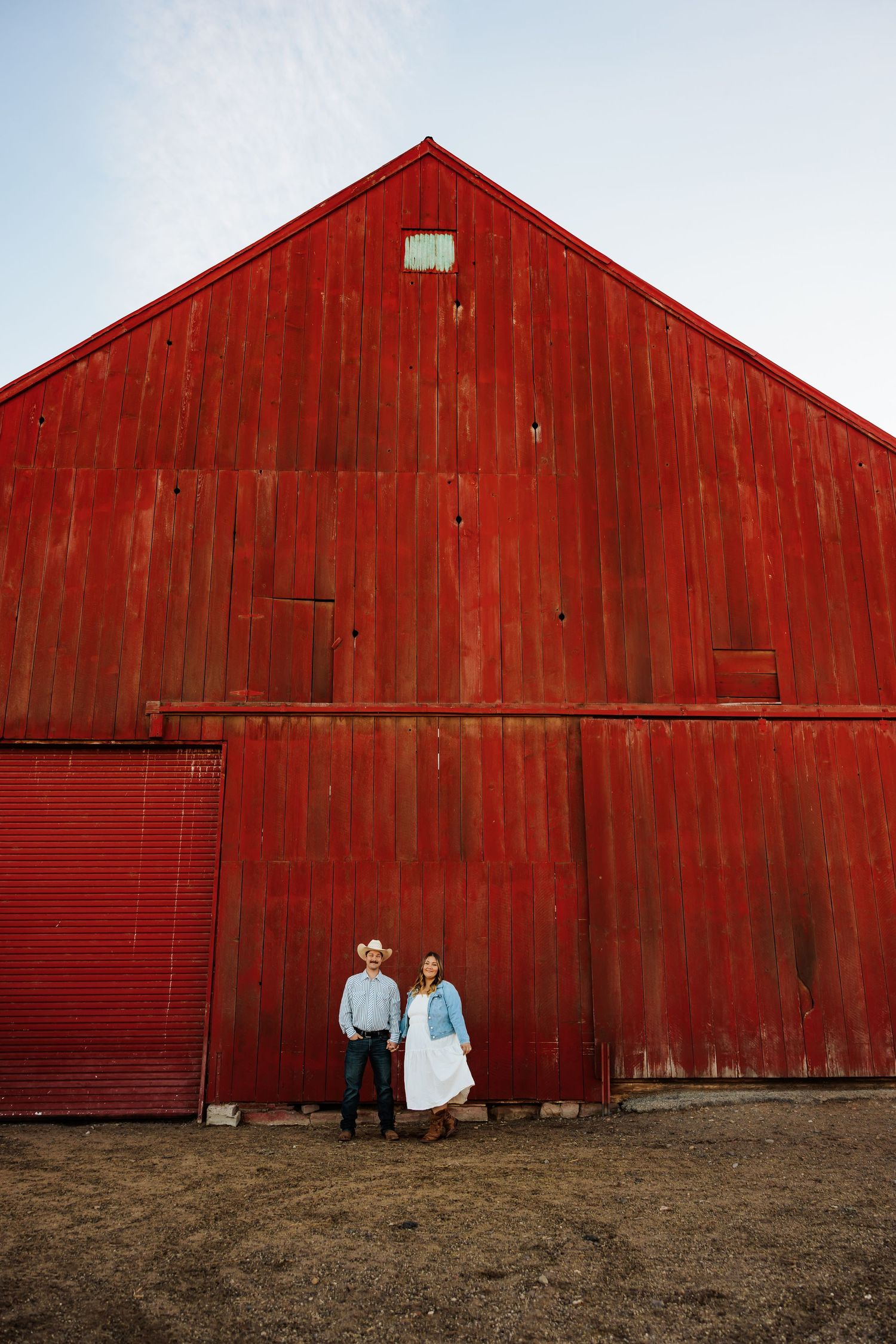 A couple stands against a large red barn in countryside setting.