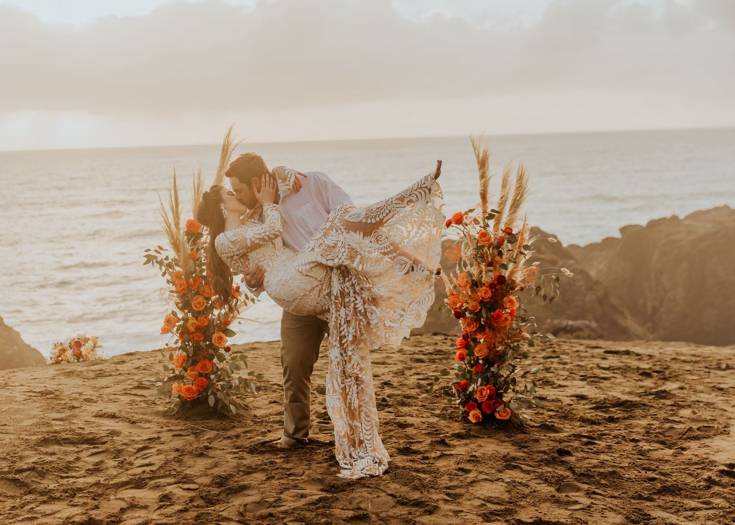 A romantic beachside wedding ceremony at sunset with floral arrangements in warm orange and rust tones framing the couple.