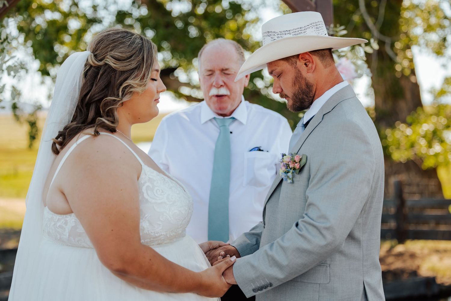 Bride and groom hold hands during outdoor wedding ceremony while officiant in suit and tie stands behind them.