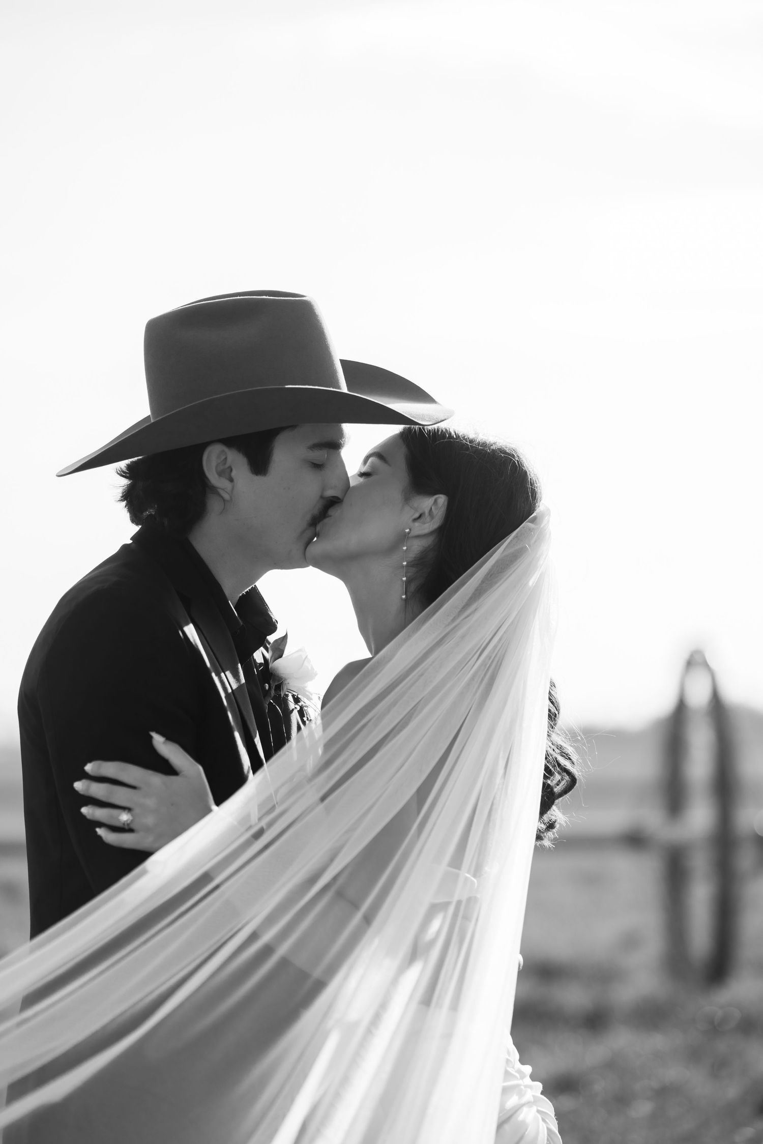Black and white photo of a couple kissing, groom in cowboy hat, bride in veil, romantic western wedding portrait.
