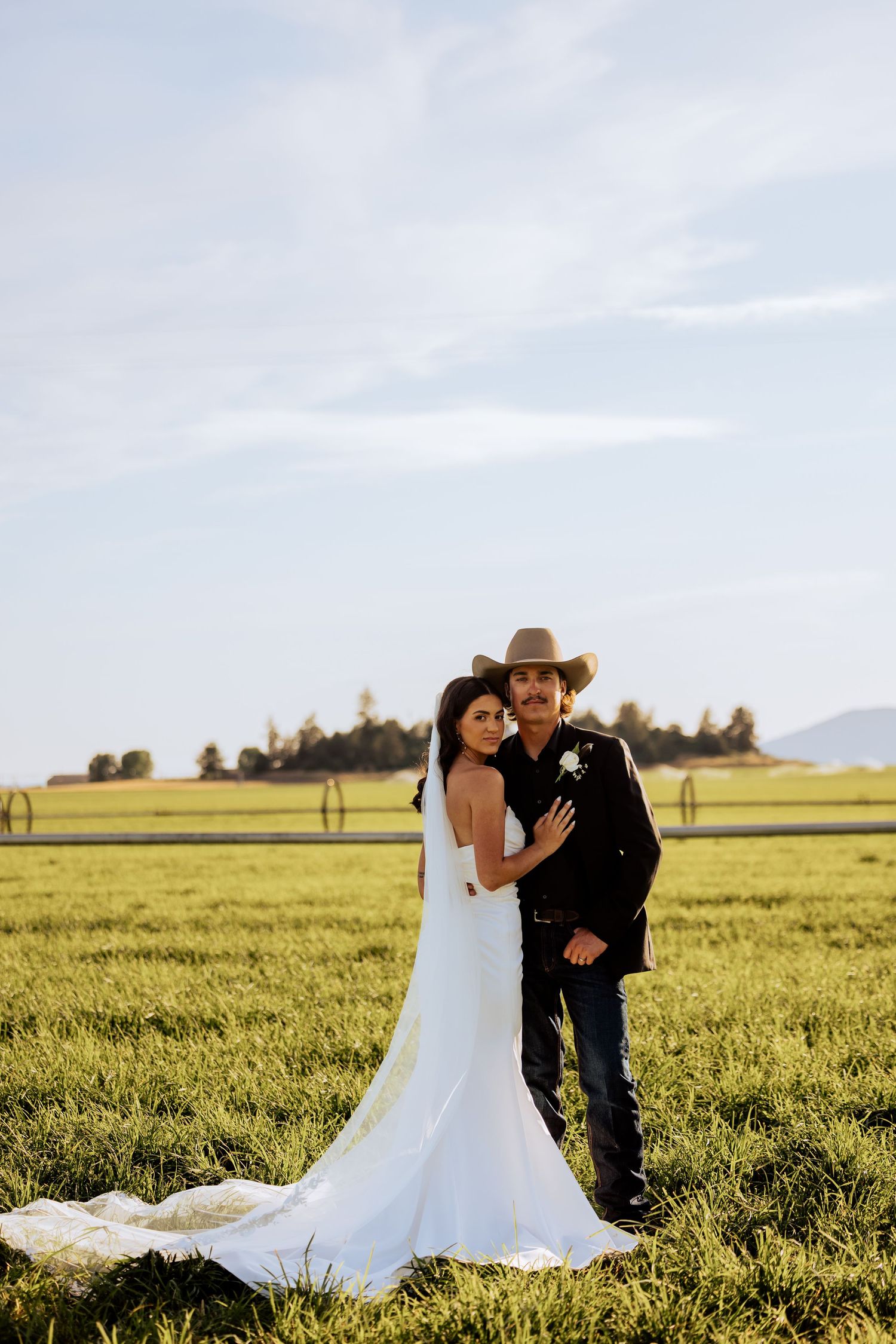 Bride in white gown and groom in western attire pose together in a golden field.