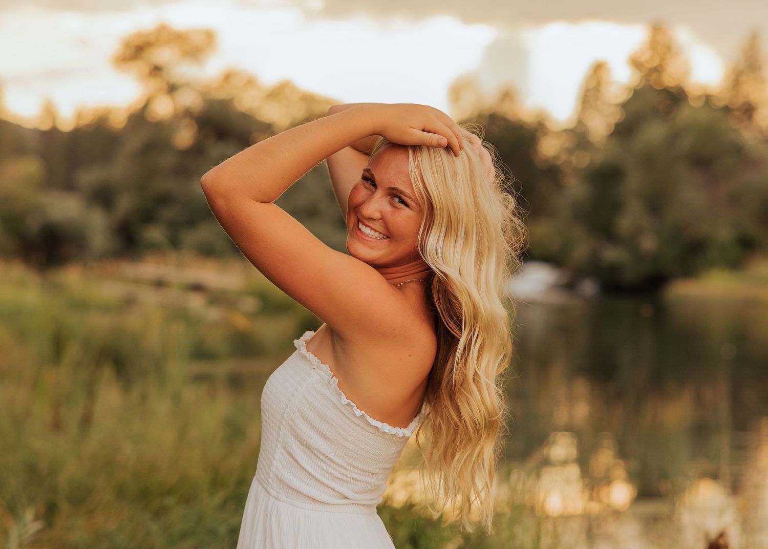 Person in white dress poses against golden sunset by lake.