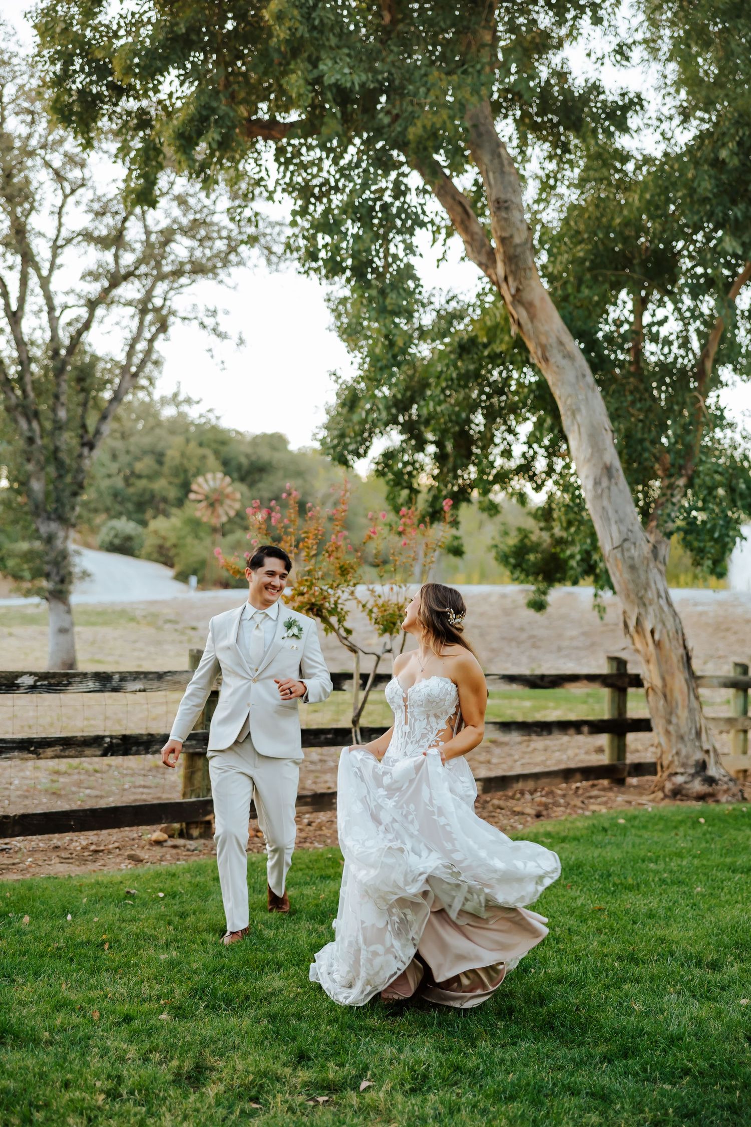 A bride and groom walk hand in hand on lush green grass beneath a tree at an outdoor wedding venue.