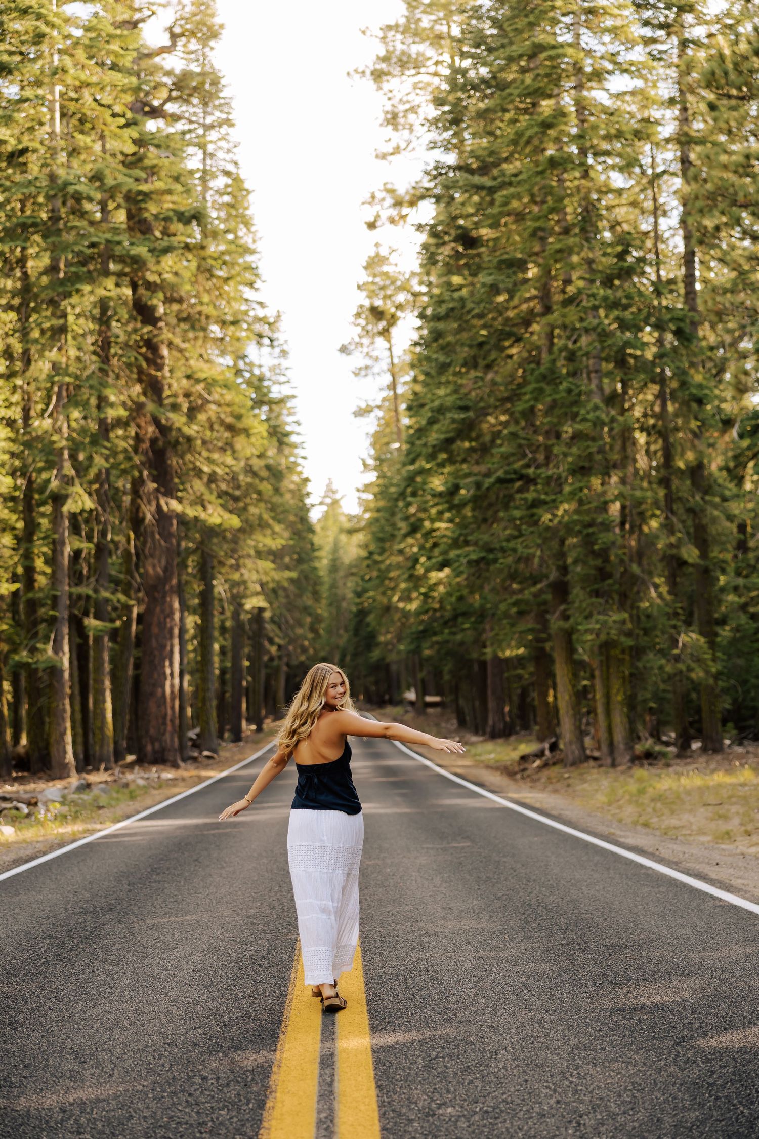 Woman in black top and white pants walks down a tree-lined road with arms outstretched.